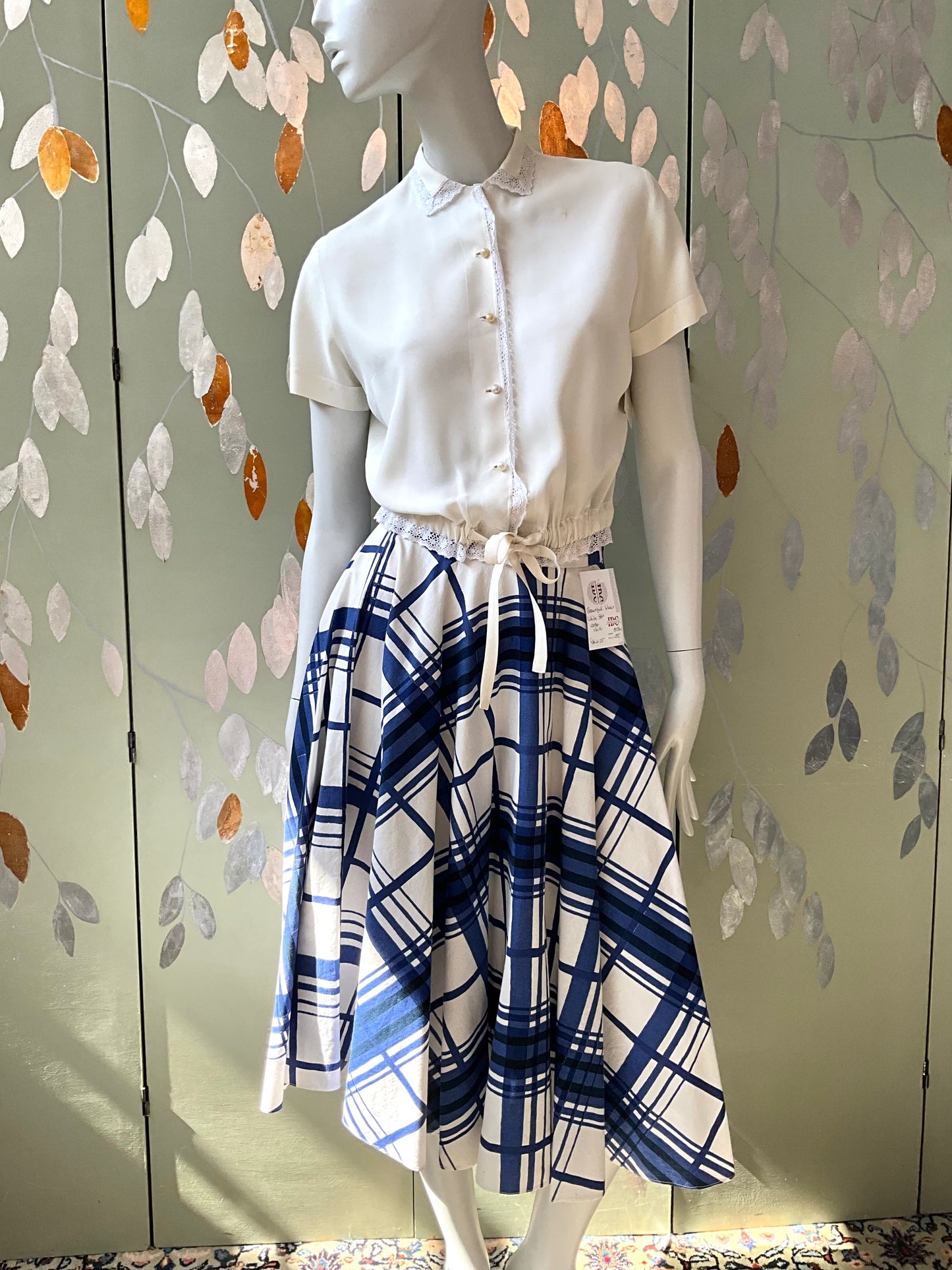 Mannequin wearing a white blouse and blue plaid skirt against a decorative wall.