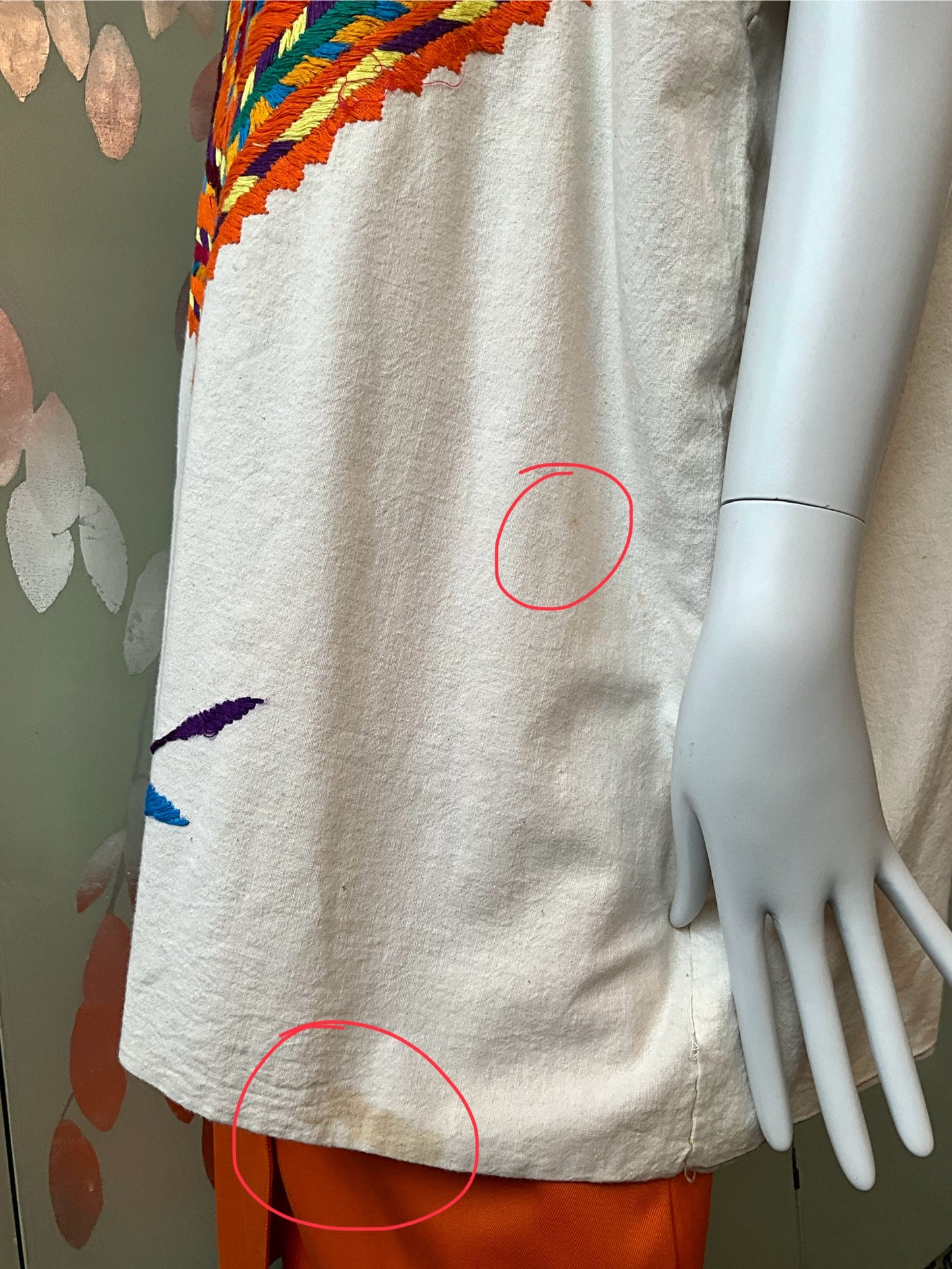 Mannequin wearing a beige garment with colorful embroidery, with a floral pattern in the background.