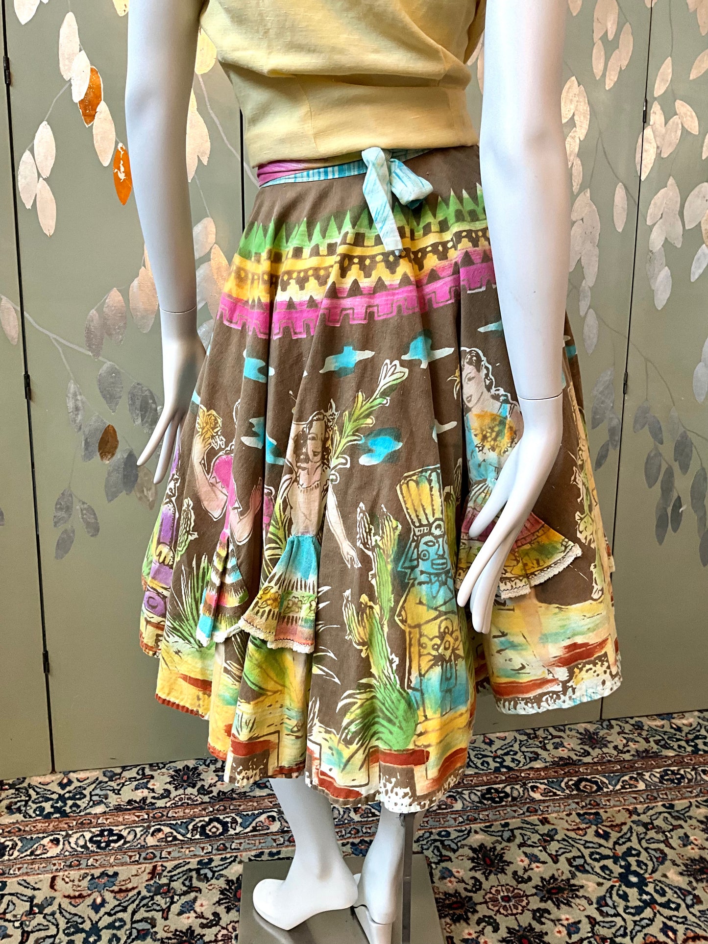 Mannequin wearing a colorful skirt with a pattern of people on a floral-patterned wall.