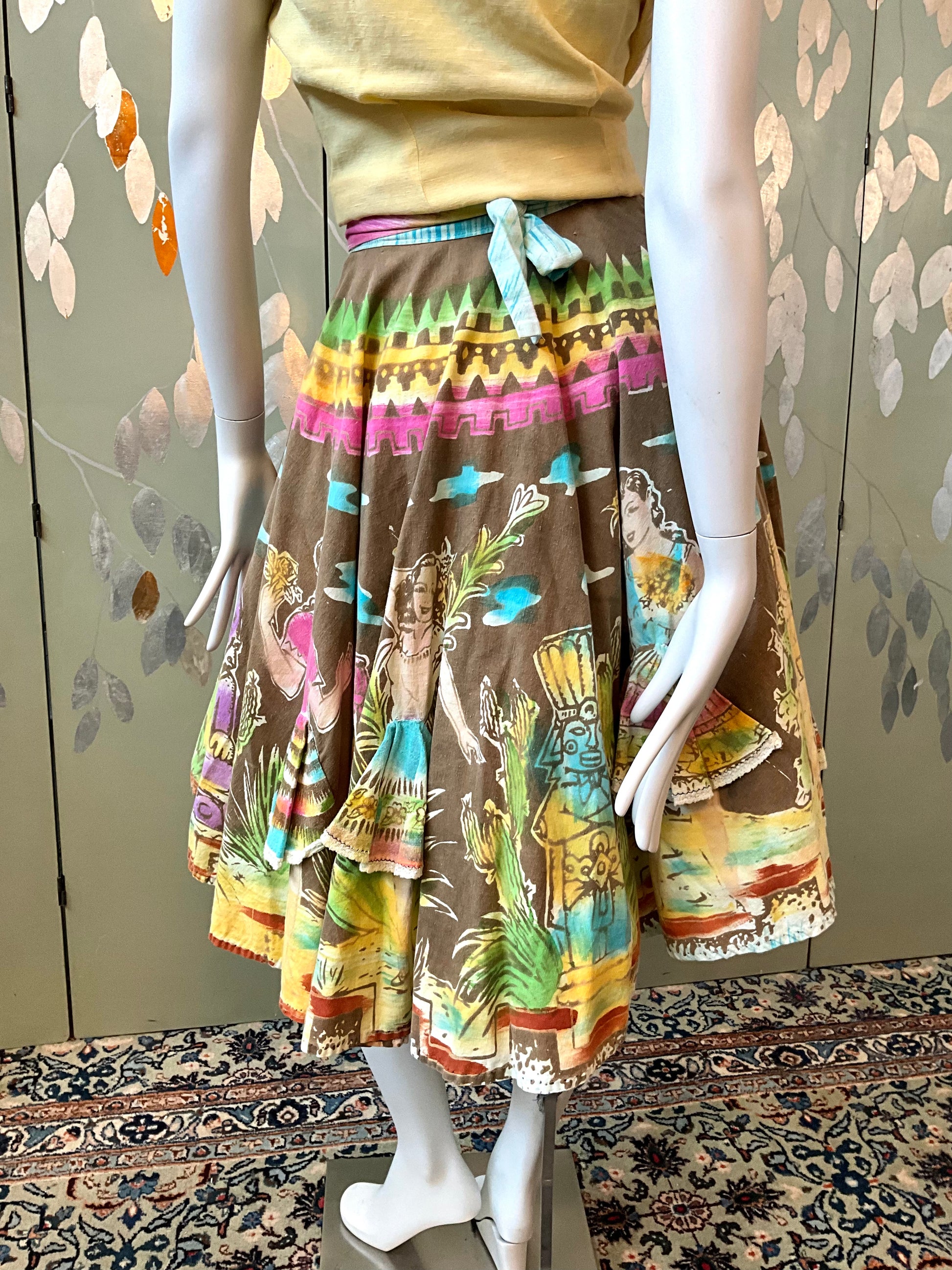Mannequin wearing a colorful skirt with a pattern of people on a floral-patterned wall.