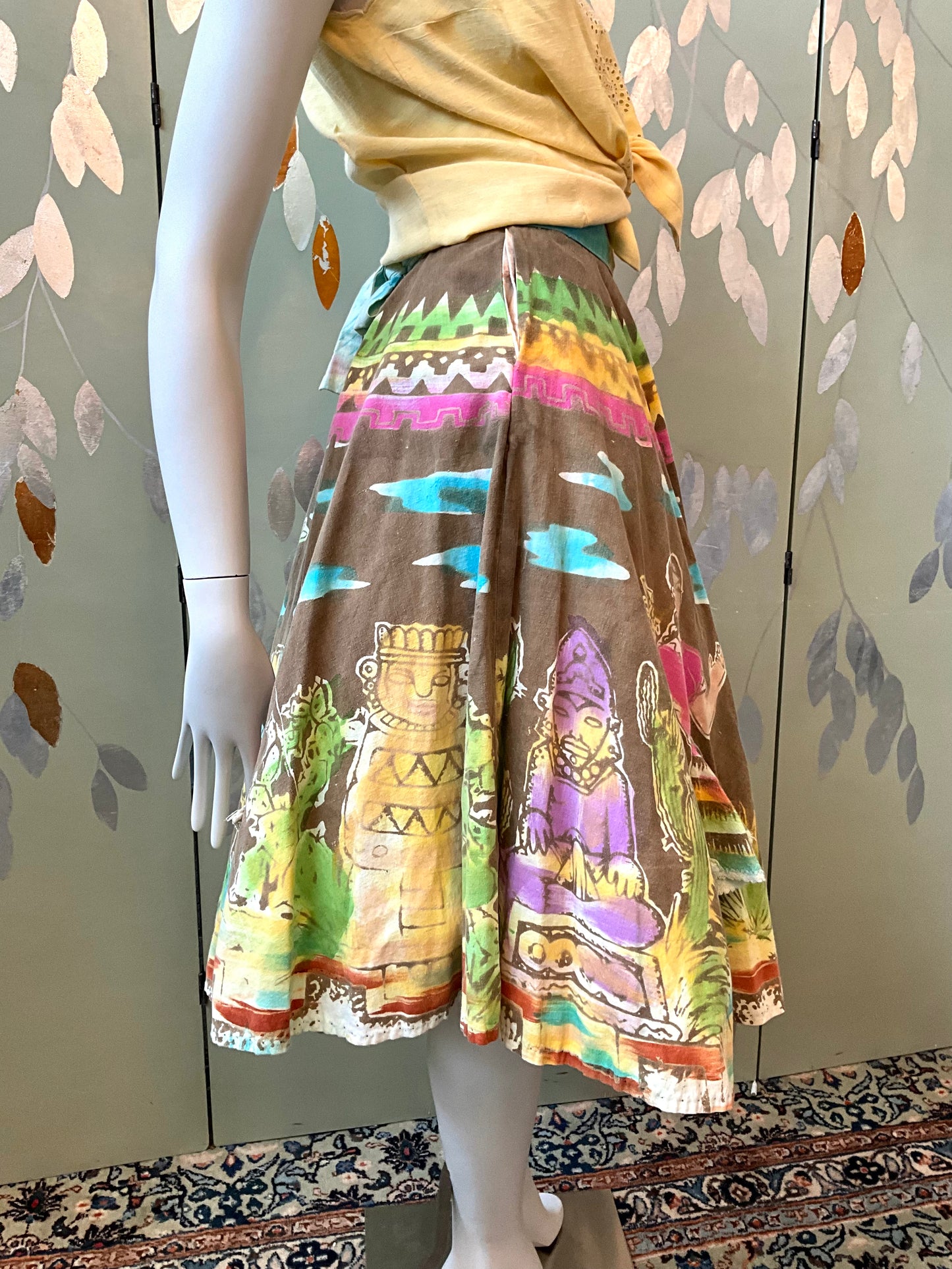 Mannequin wearing a colorful skirt with tribal designs against a patterned wall.