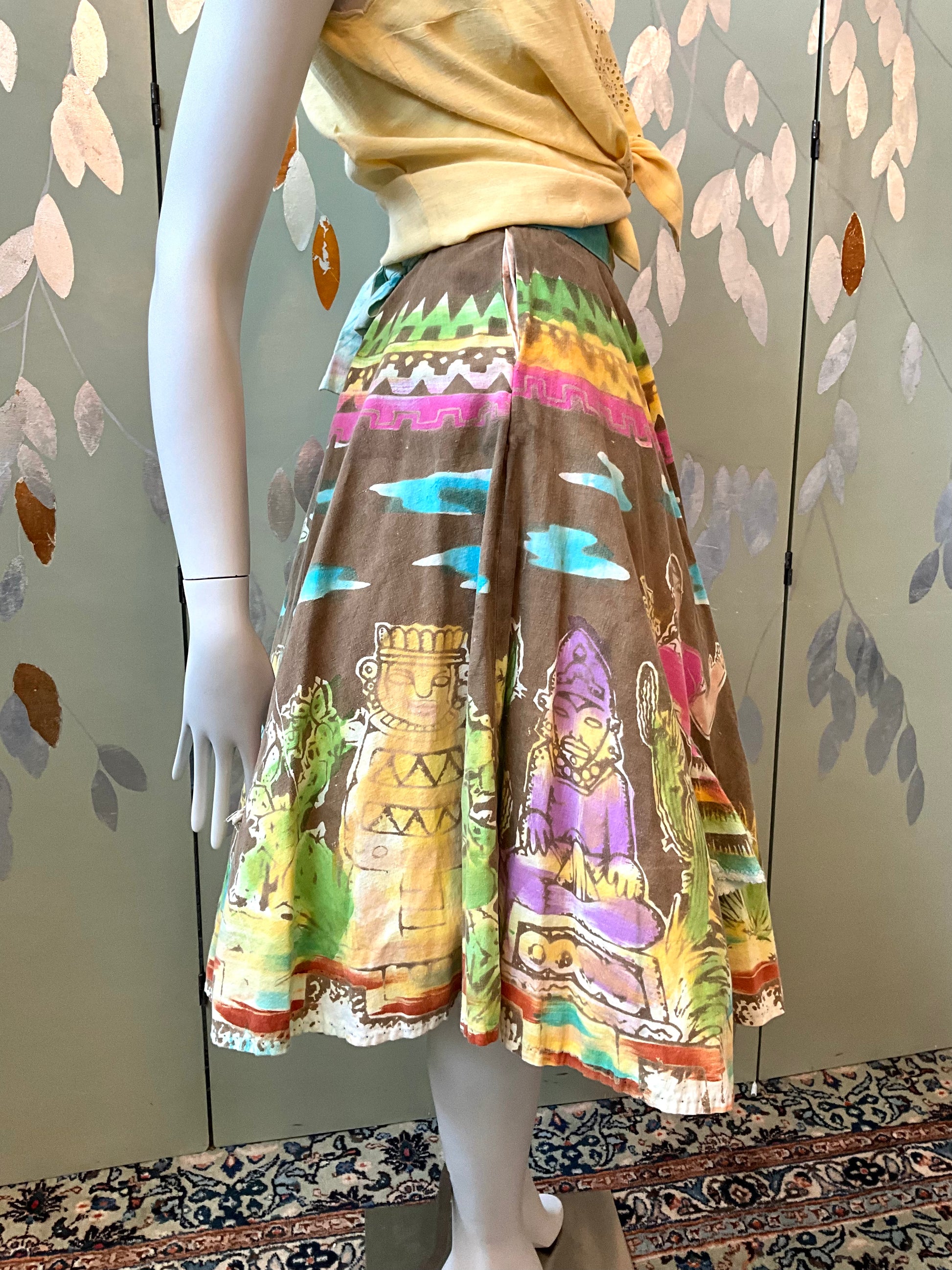 Mannequin wearing a colorful skirt with tribal designs against a patterned wall.
