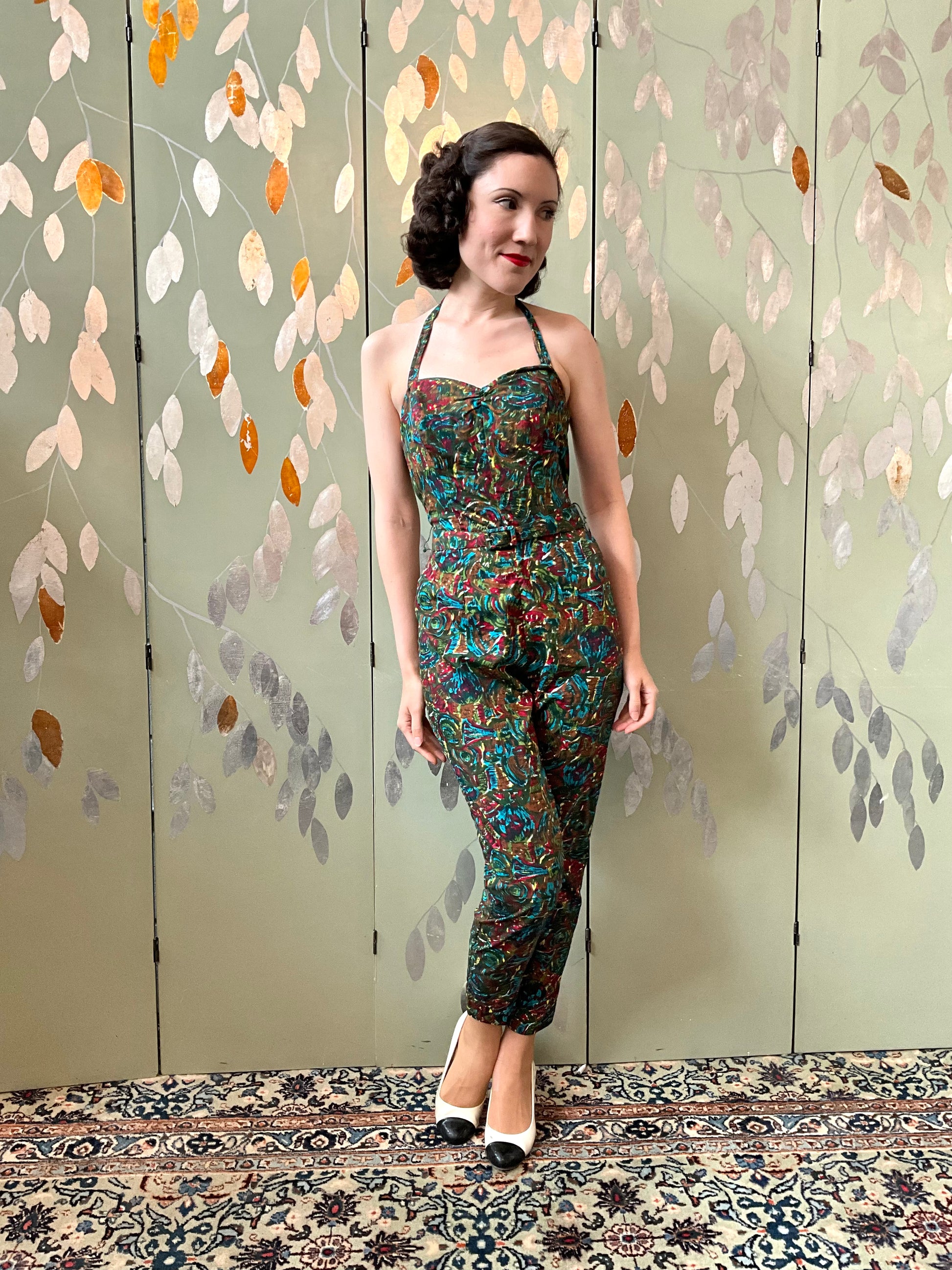Woman in a patterned jumpsuit standing against a decorative wall with leaf patterns.