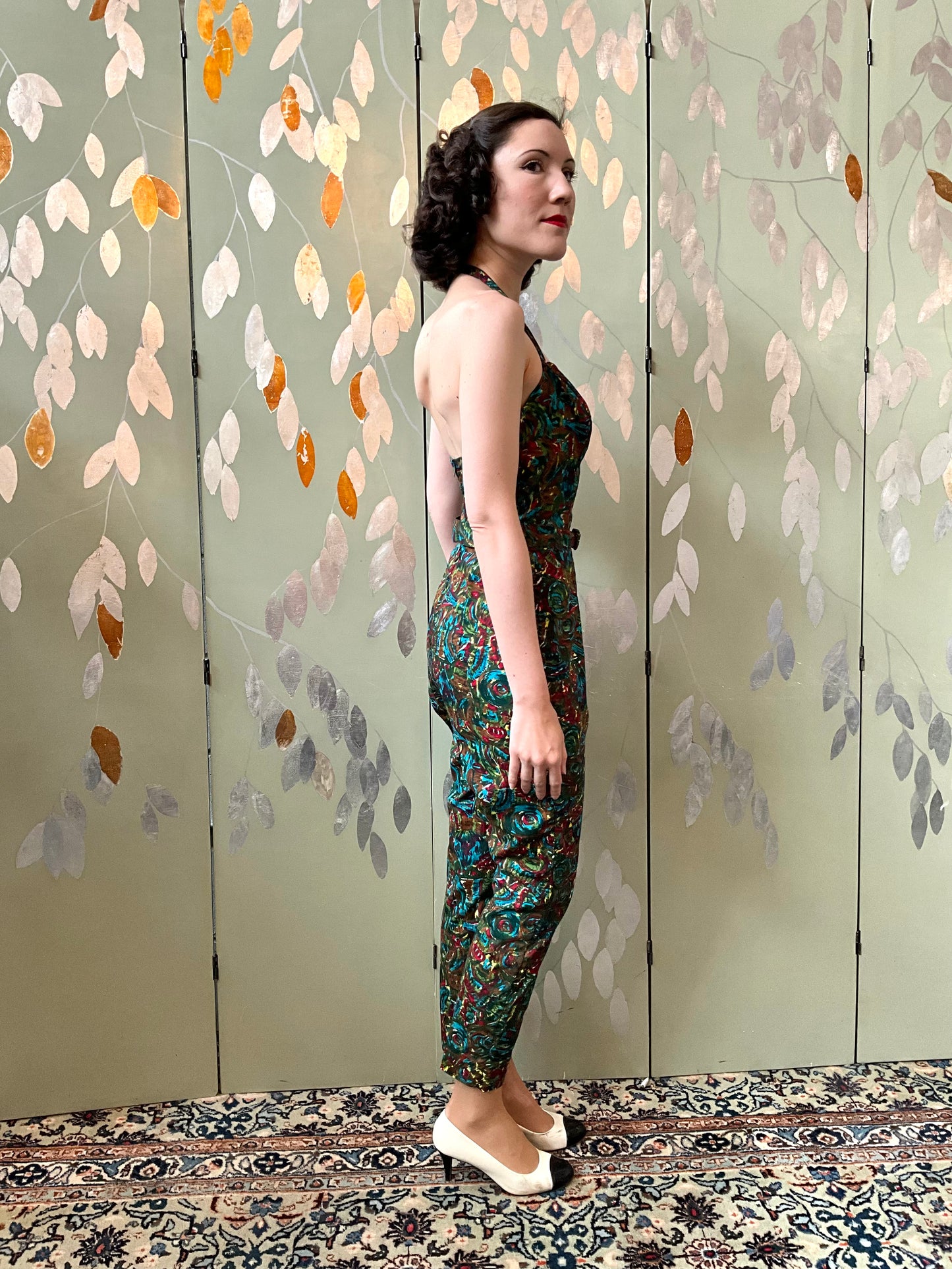 Woman in a patterned jumpsuit standing against a decorative wall with leaf patterns.