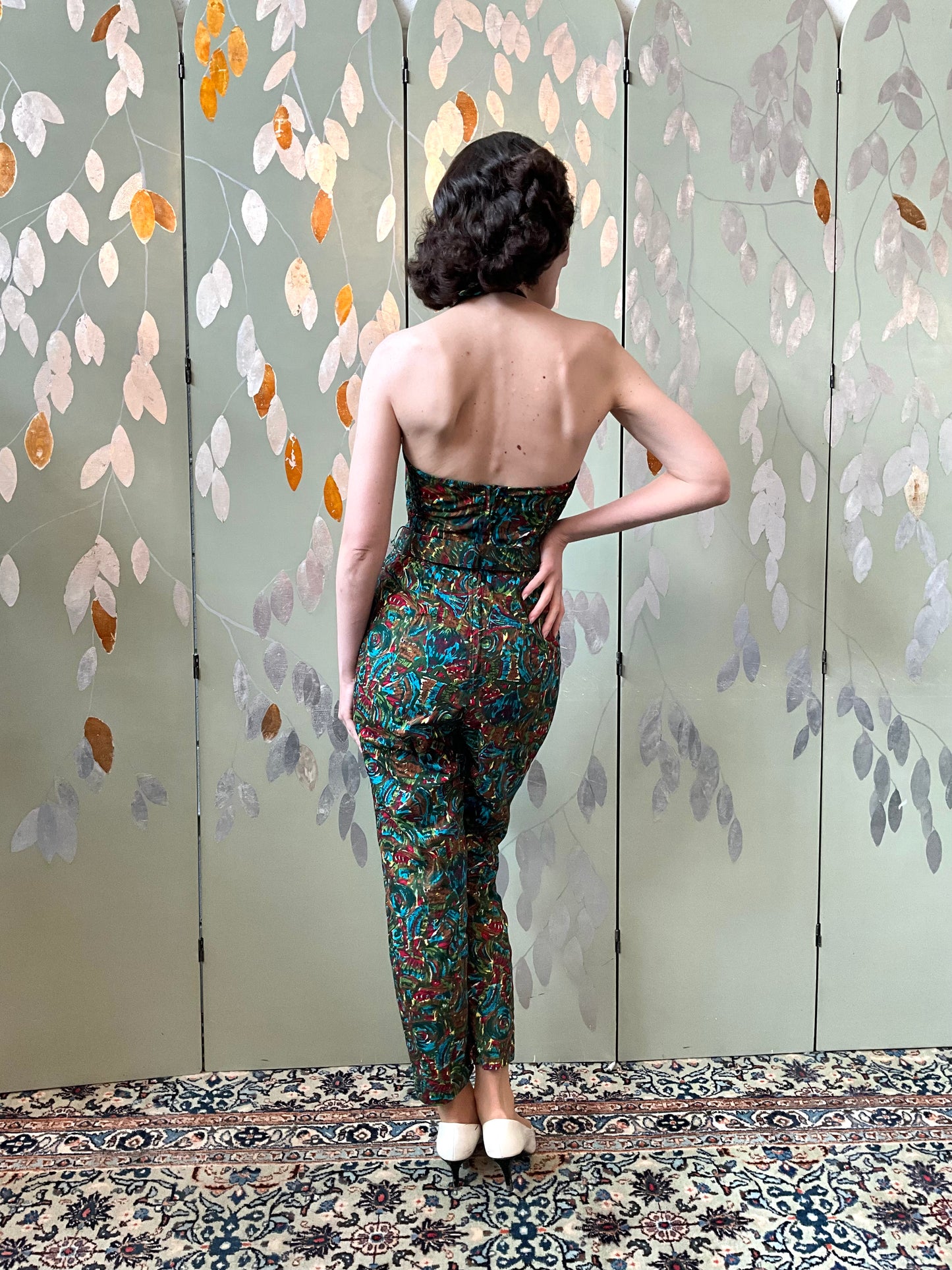 Woman in a patterned jumpsuit standing in front of a decorative wall with leaf patterns.