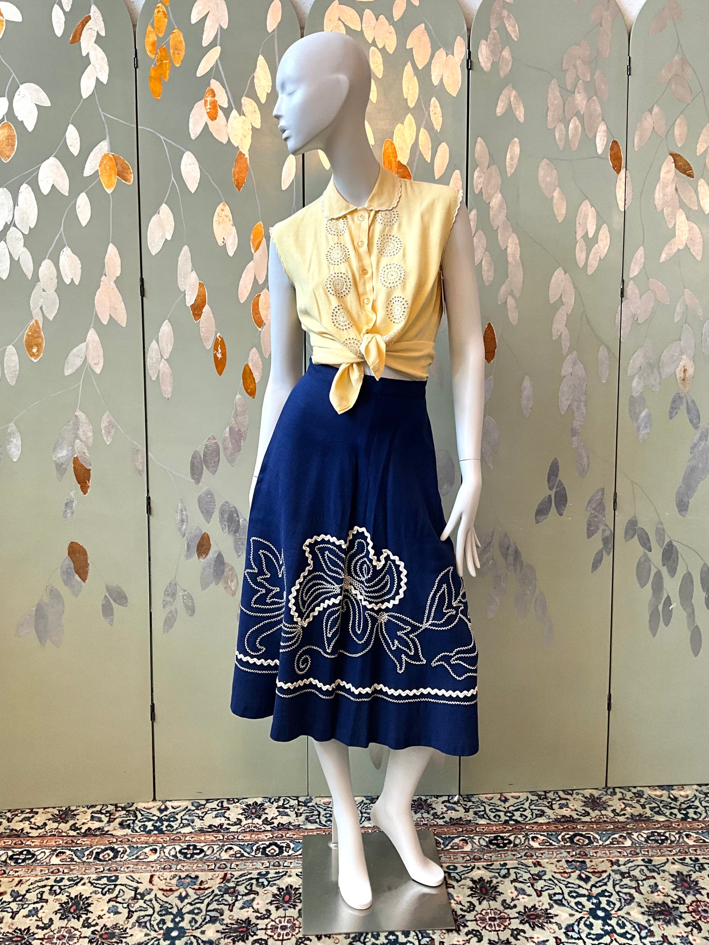 Mannequin wearing a yellow top and blue skirt with floral embroidery against a decorative wall.