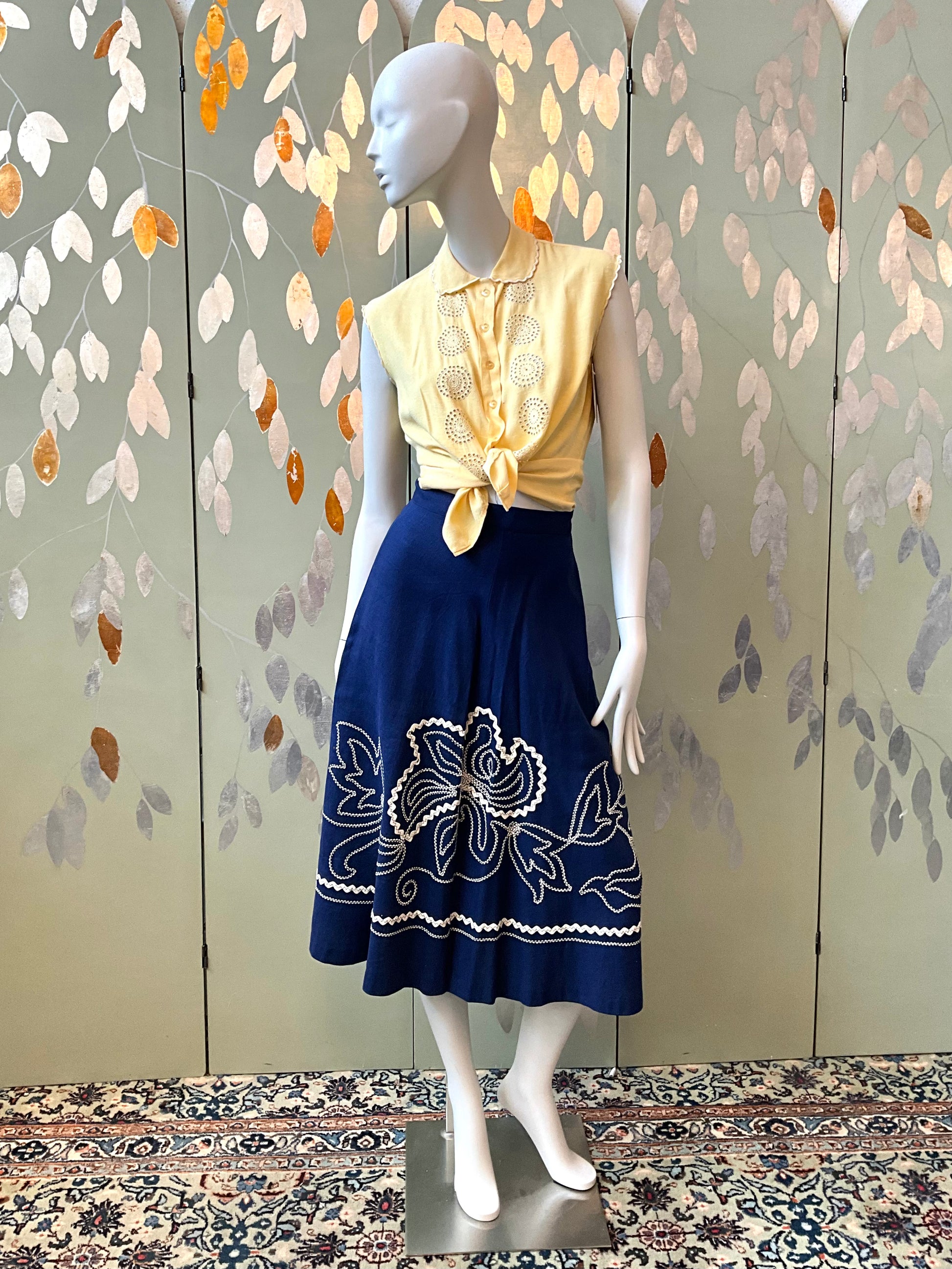 Mannequin wearing a yellow top and blue skirt with floral embroidery against a decorative wall.