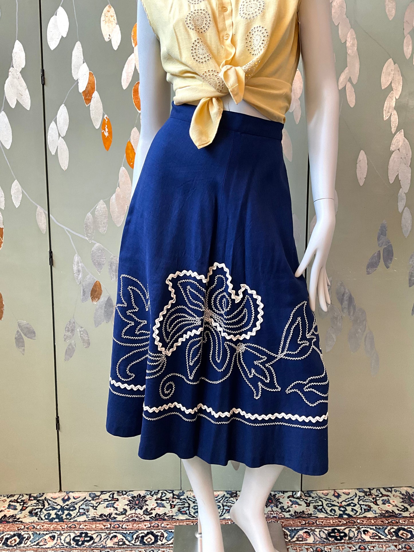 Mannequin wearing a blue skirt with white floral embroidery against a decorative wall.