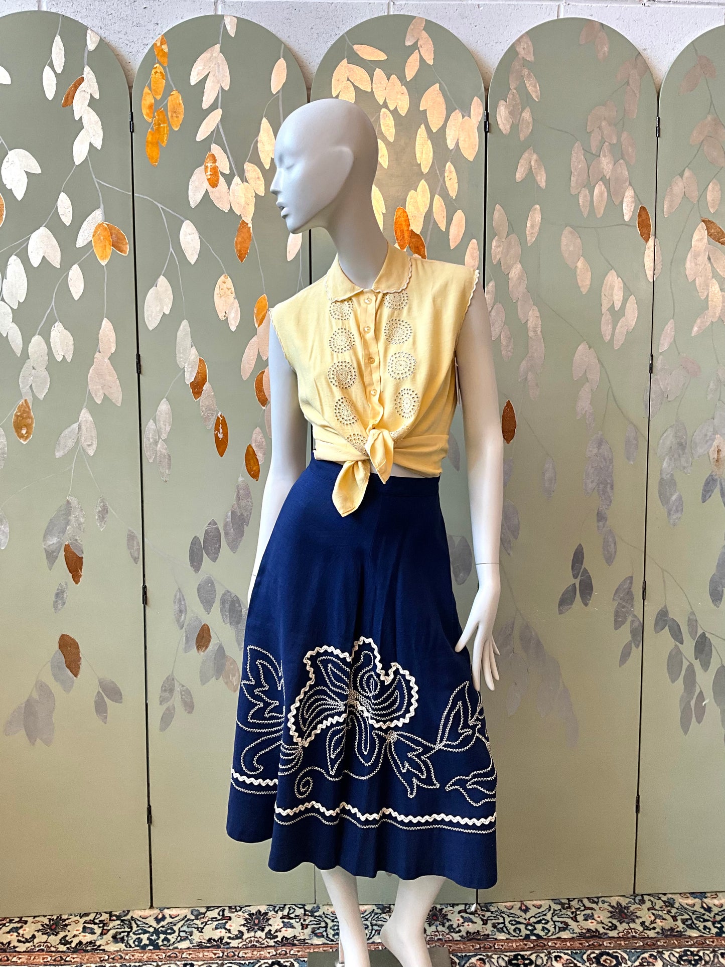 Mannequin wearing a yellow top and blue skirt with floral embroidery against a decorative wall.