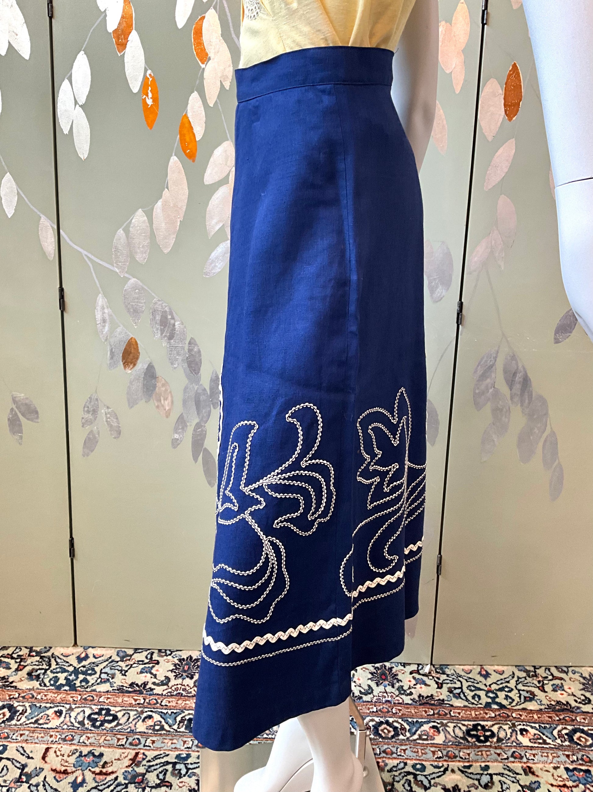 Blue skirt with white embroidery on a mannequin against a floral pattern wall.