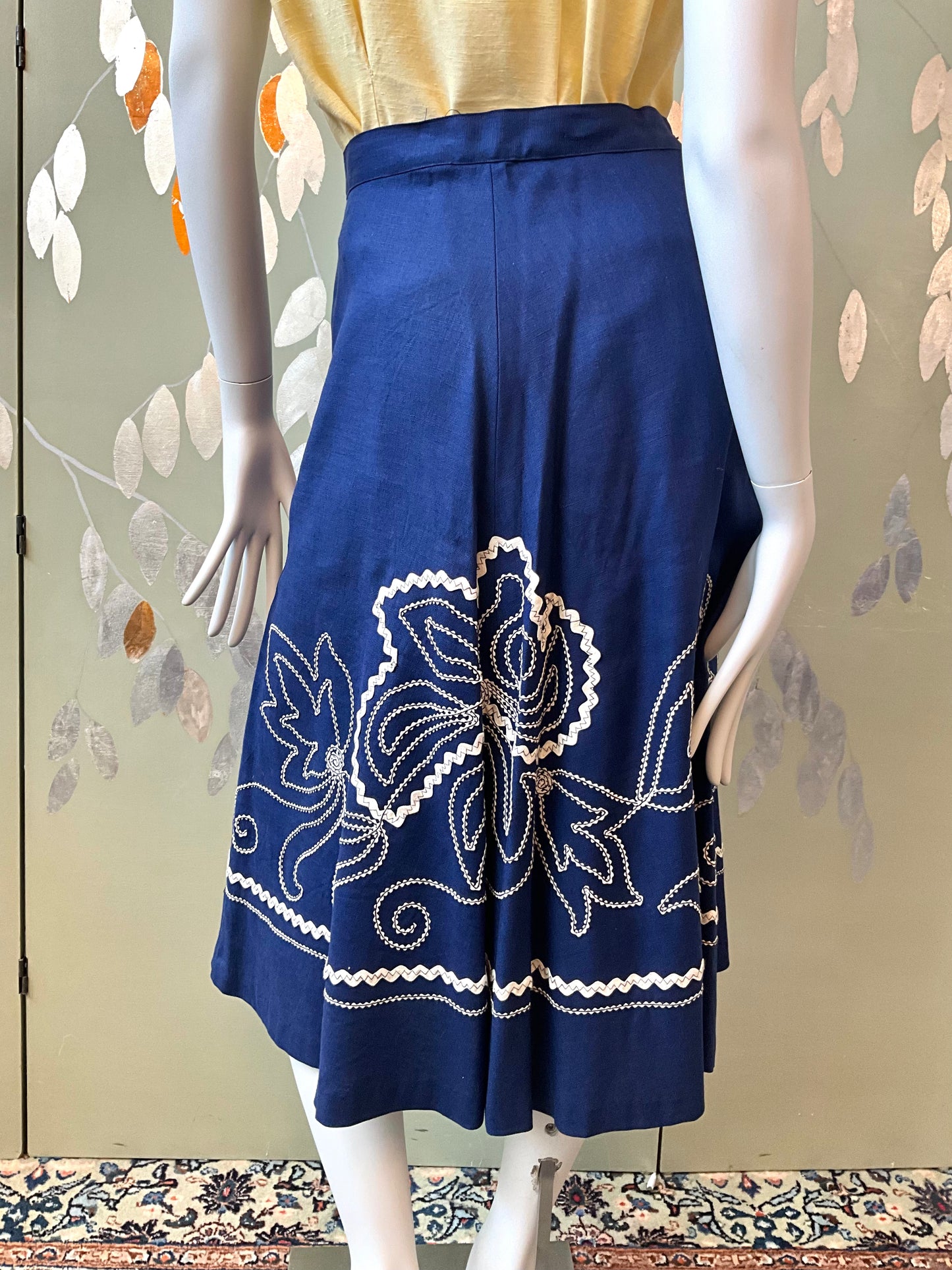 Mannequin wearing a blue skirt with white floral embroidery against a decorative wall.