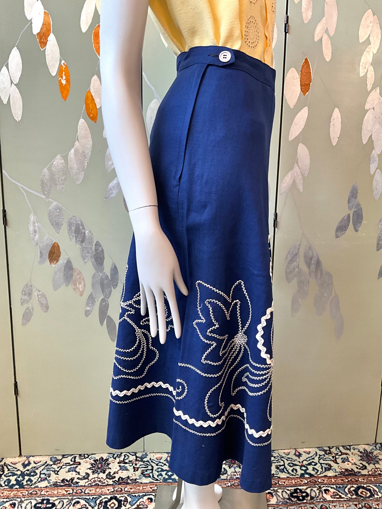 Mannequin wearing a blue skirt with white embroidery against a patterned wall.