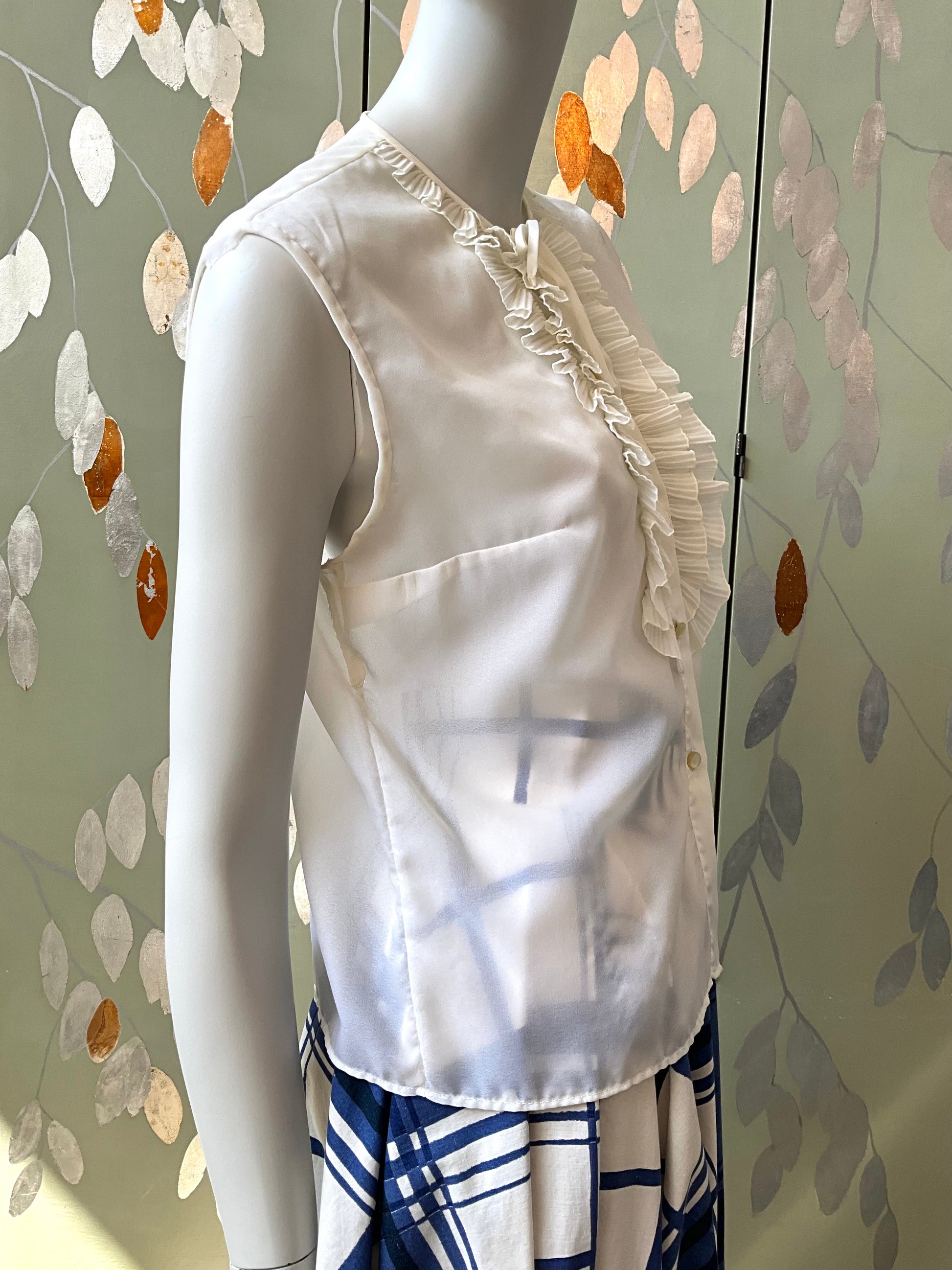 White sleeveless top with ruffled details on a mannequin against a decorative wall.