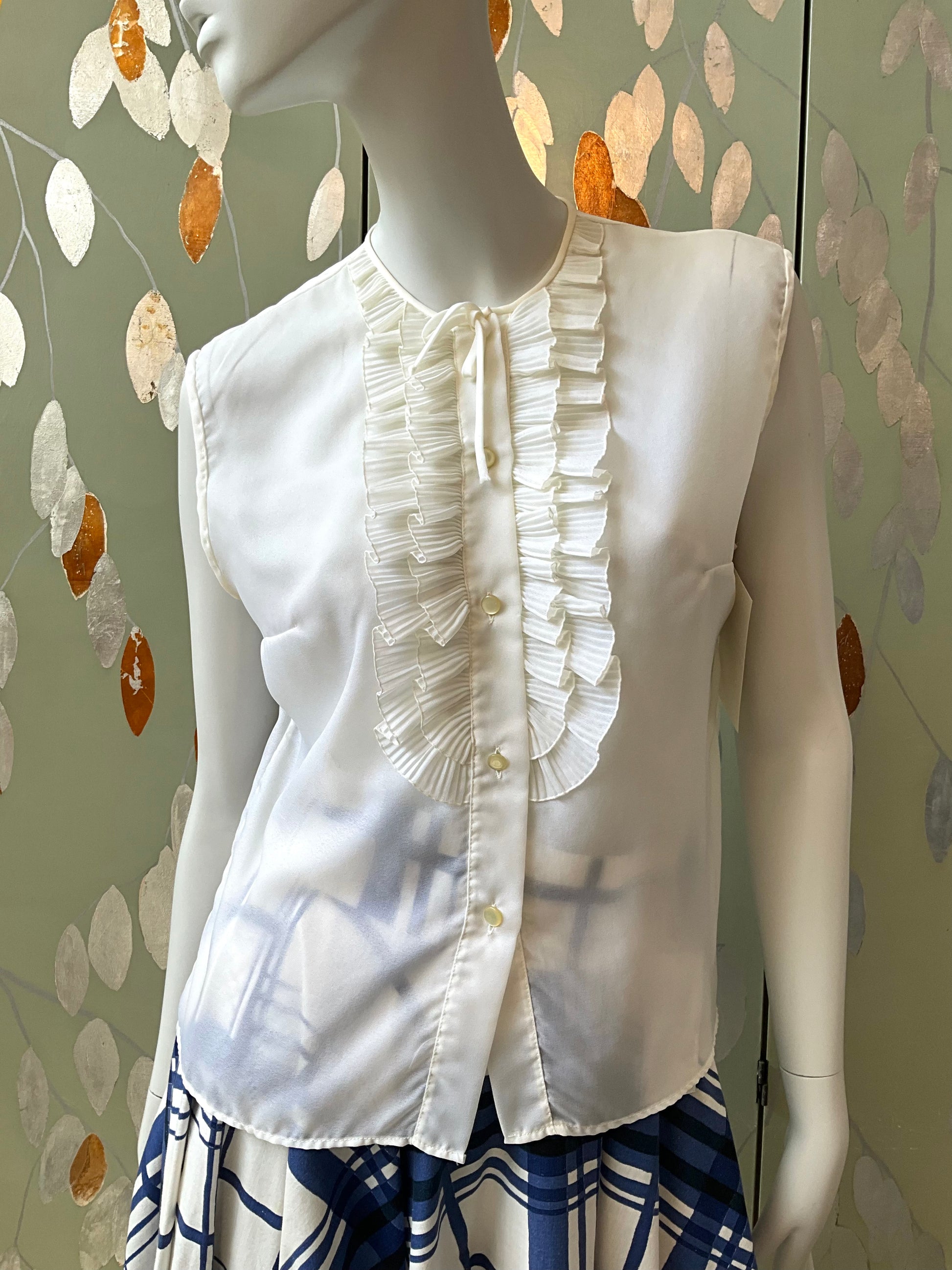 White blouse with ruffled details on a mannequin against a decorative wall.