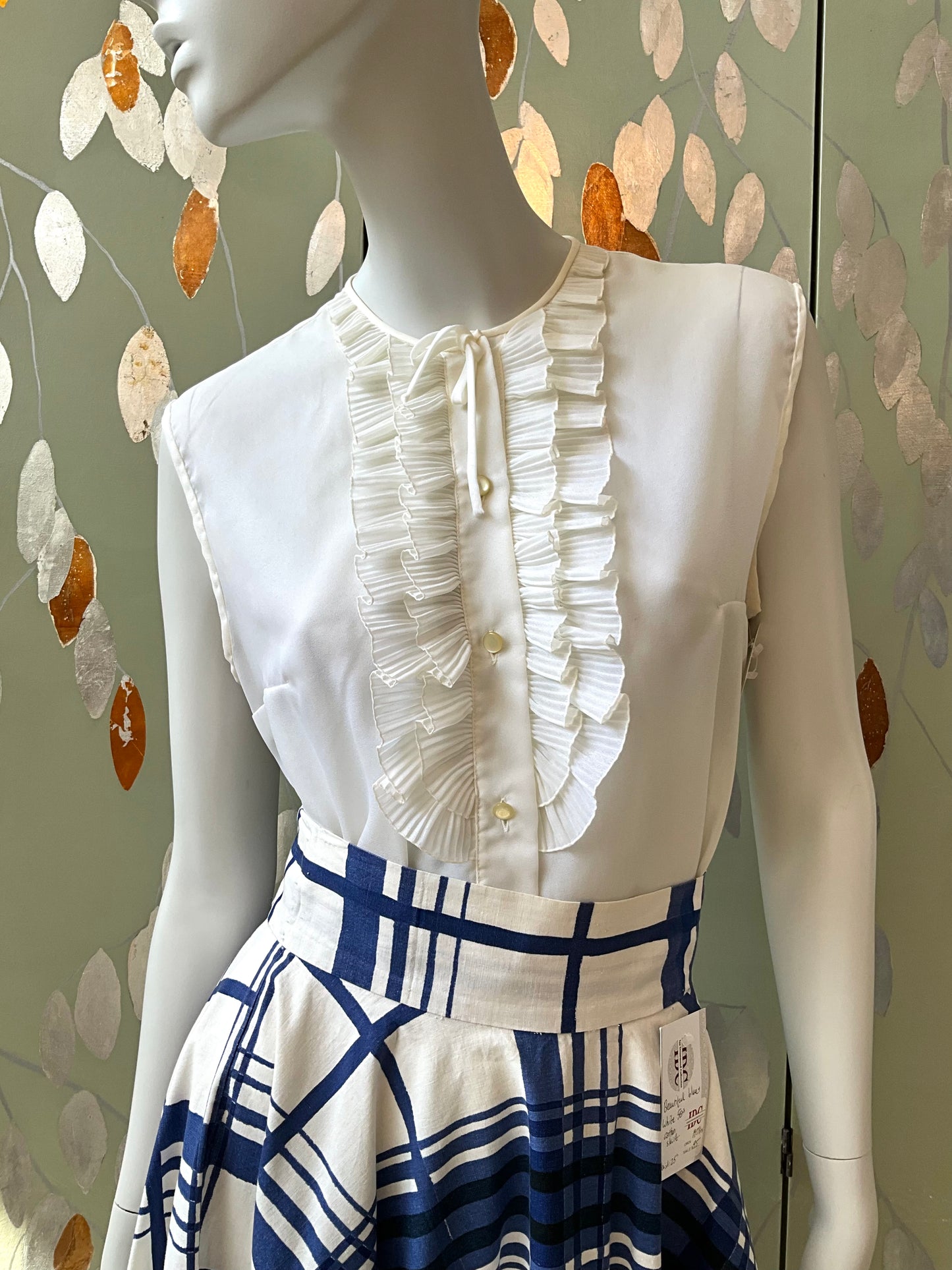 Mannequin wearing a white blouse with ruffled details and a blue and white checkered skirt against a decorative wall.