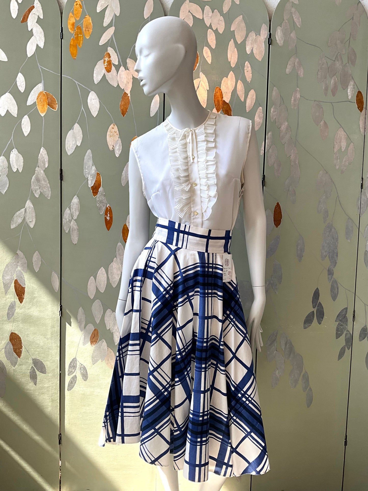 Mannequin wearing a white blouse with ruffles and a blue plaid skirt against a decorative wall.