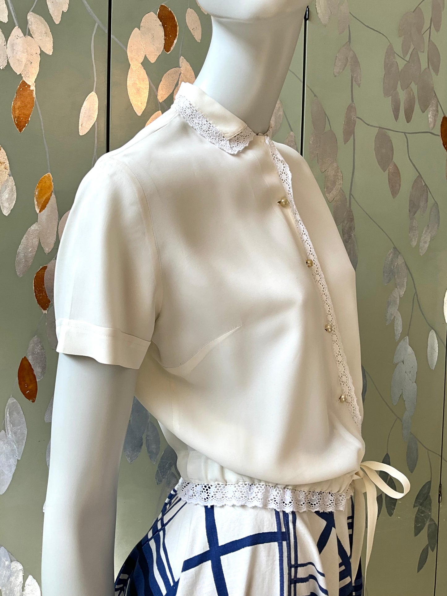 Mannequin wearing a white lace-trimmed blouse against a decorative wall.