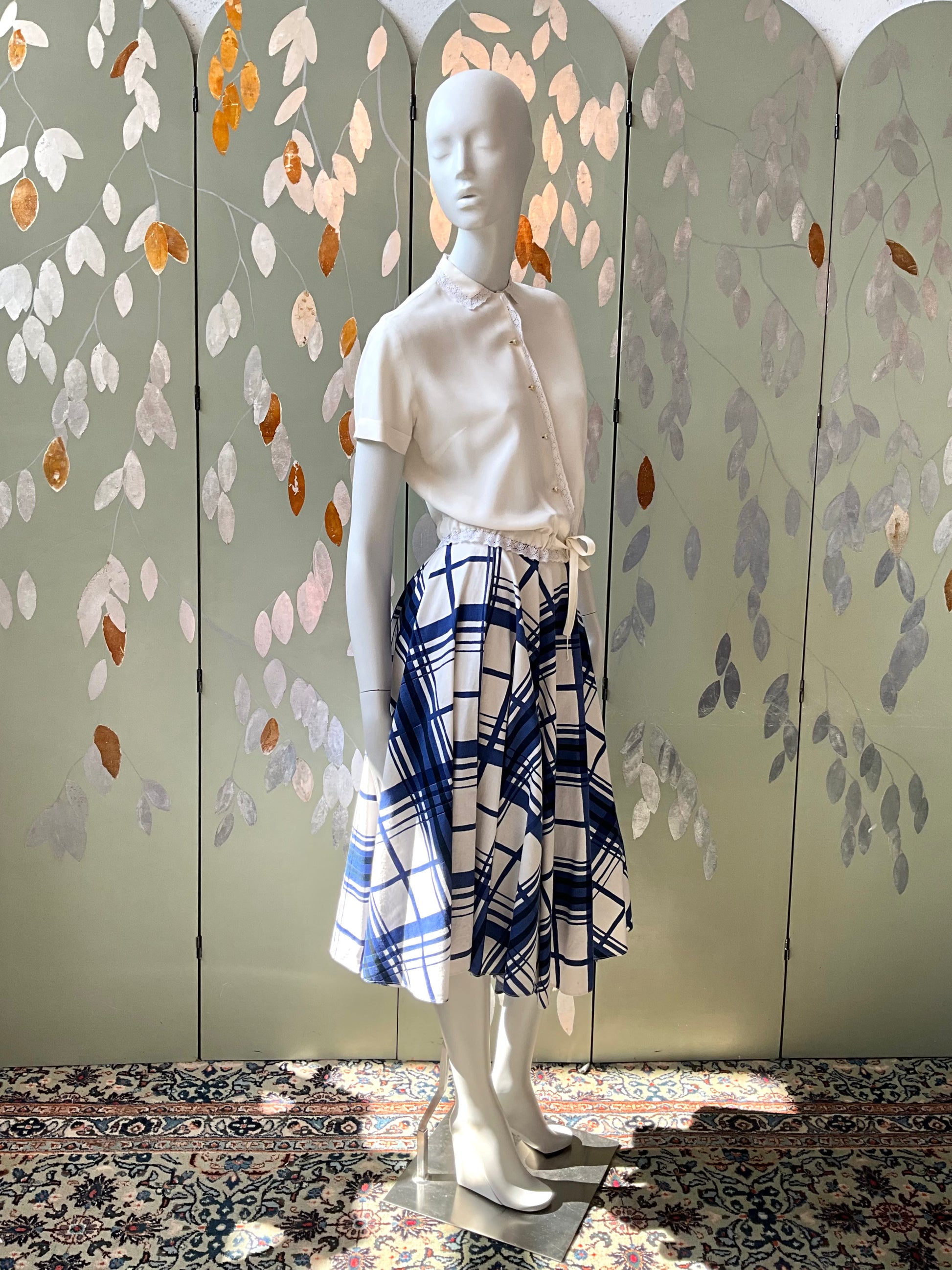 Mannequin wearing a white top and plaid skirt against a decorative wall.