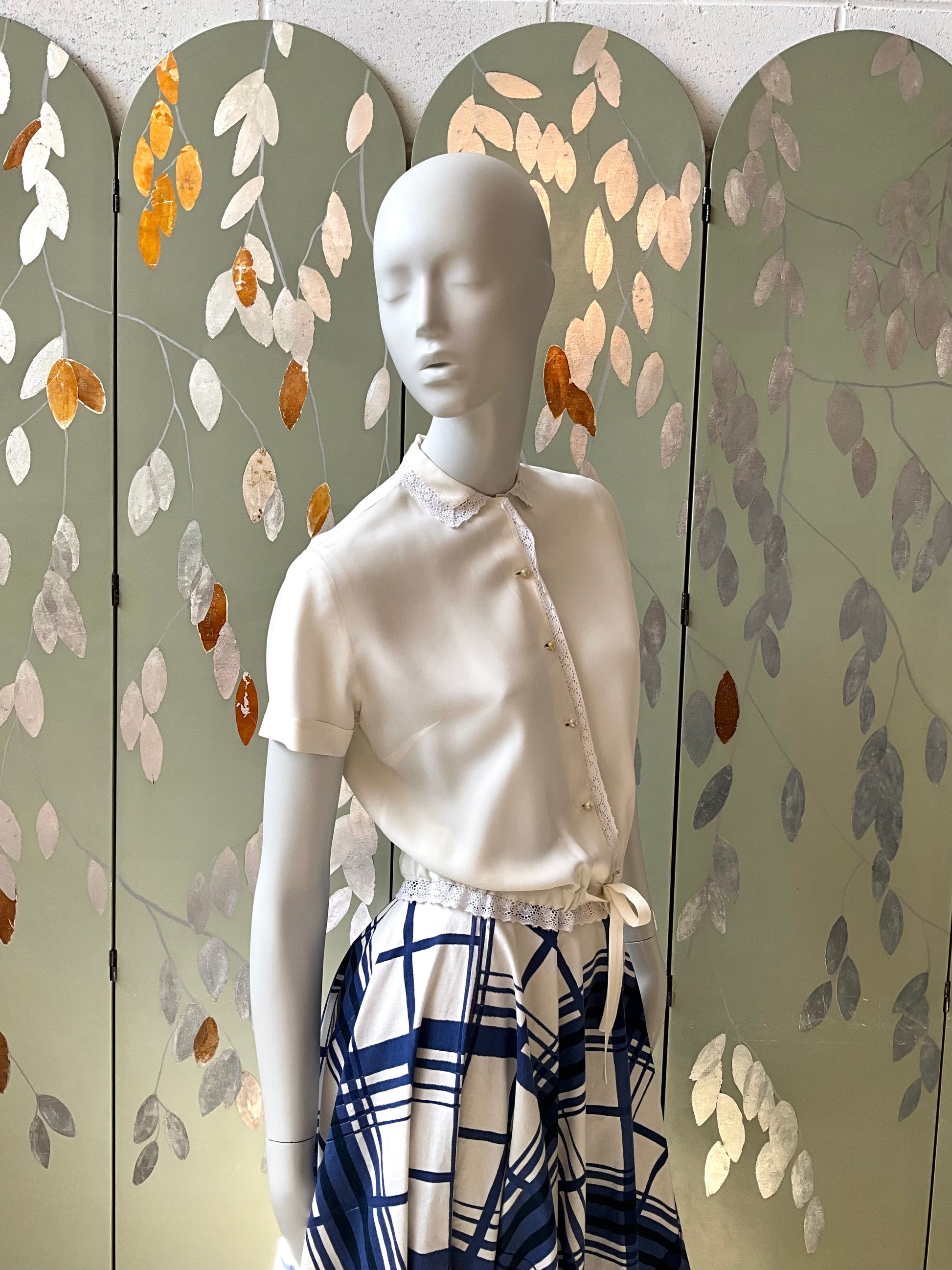 Mannequin wearing a white shirt and blue plaid skirt against a decorative screen with leaf patterns.