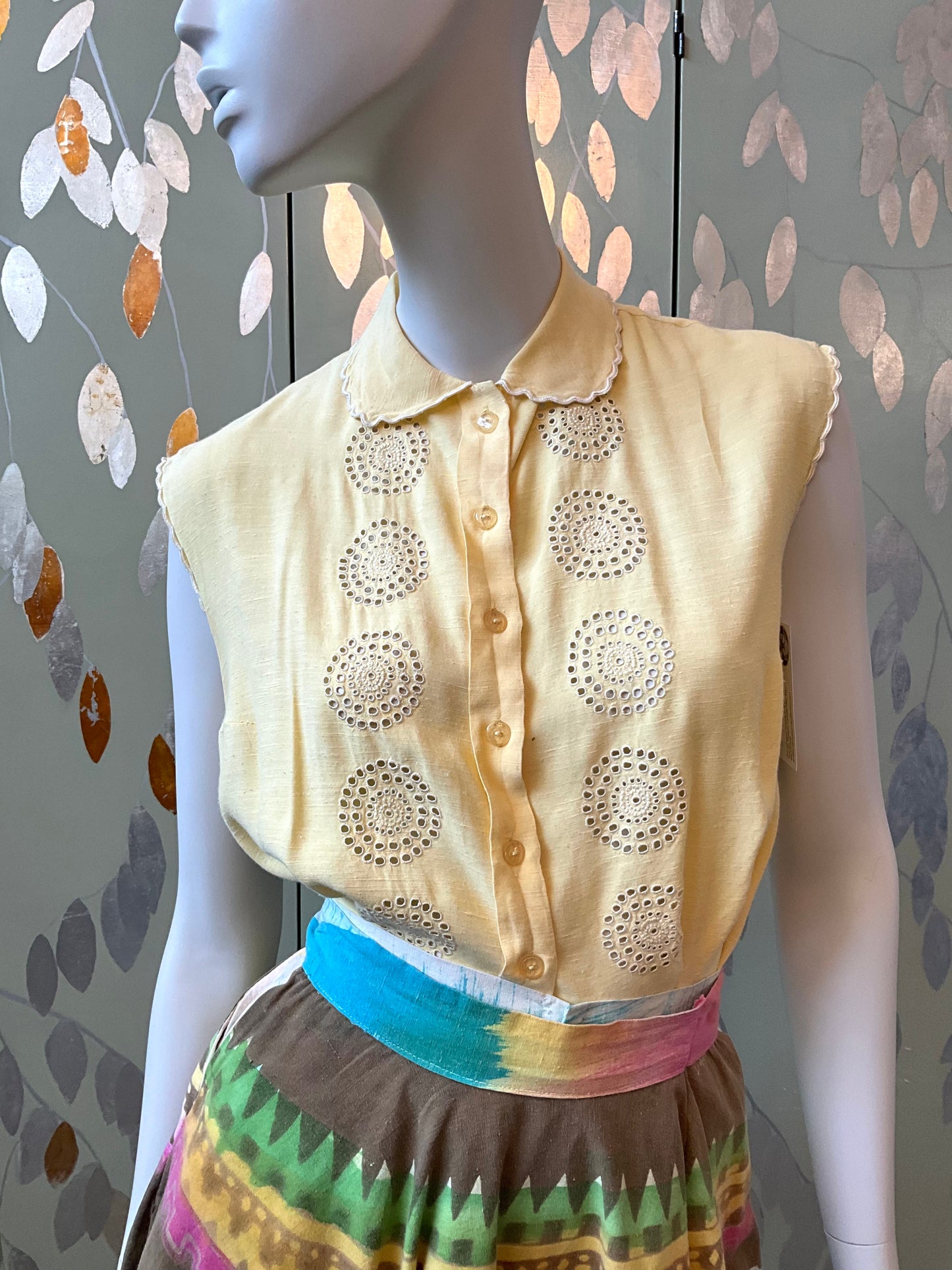 Mannequin wearing a yellow sleeveless top with decorative cut-out patterns against a patterned wall.