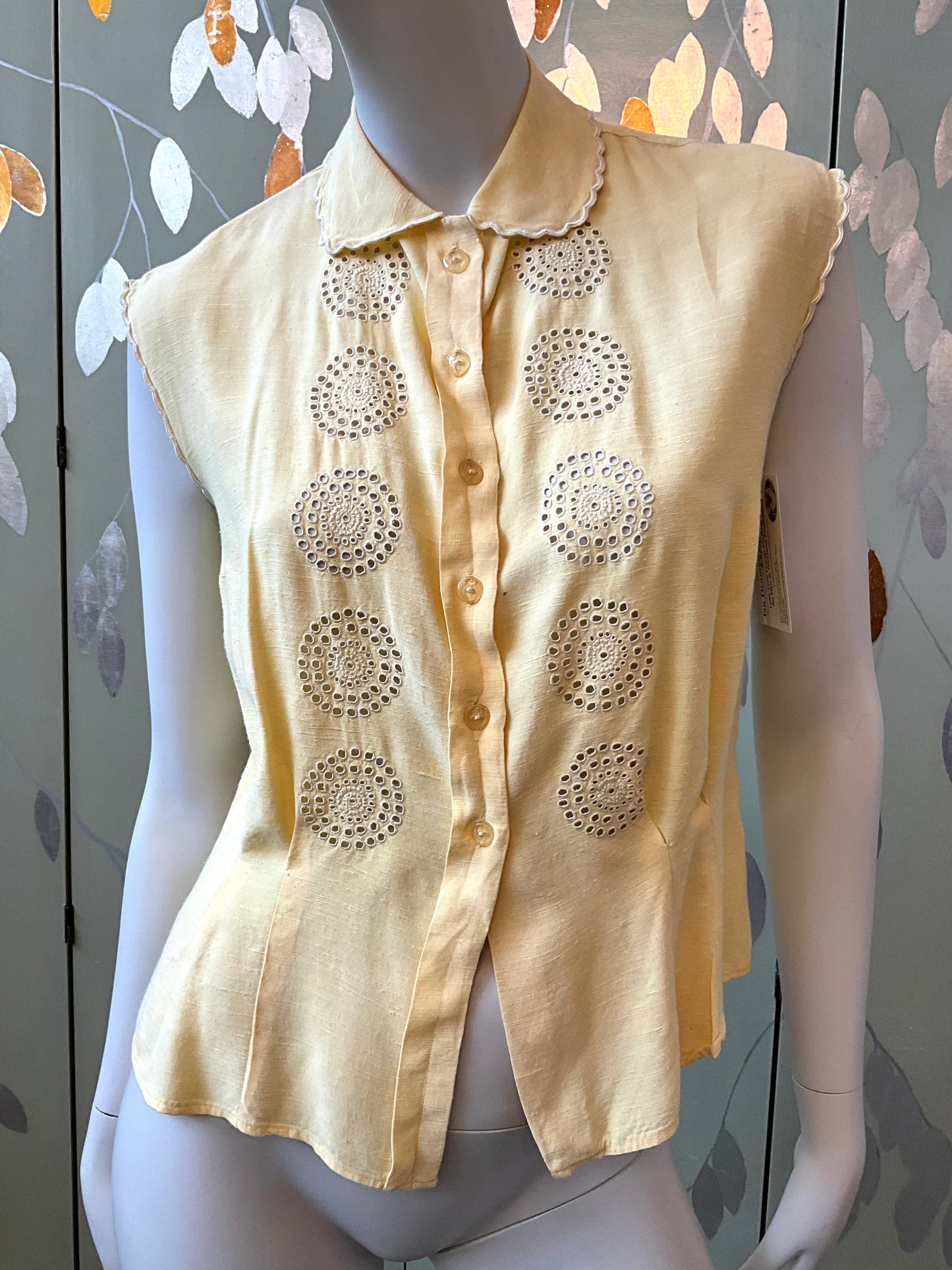 Yellow sleeveless blouse with intricate cut-out patterns on a mannequin against a floral-patterned wall.