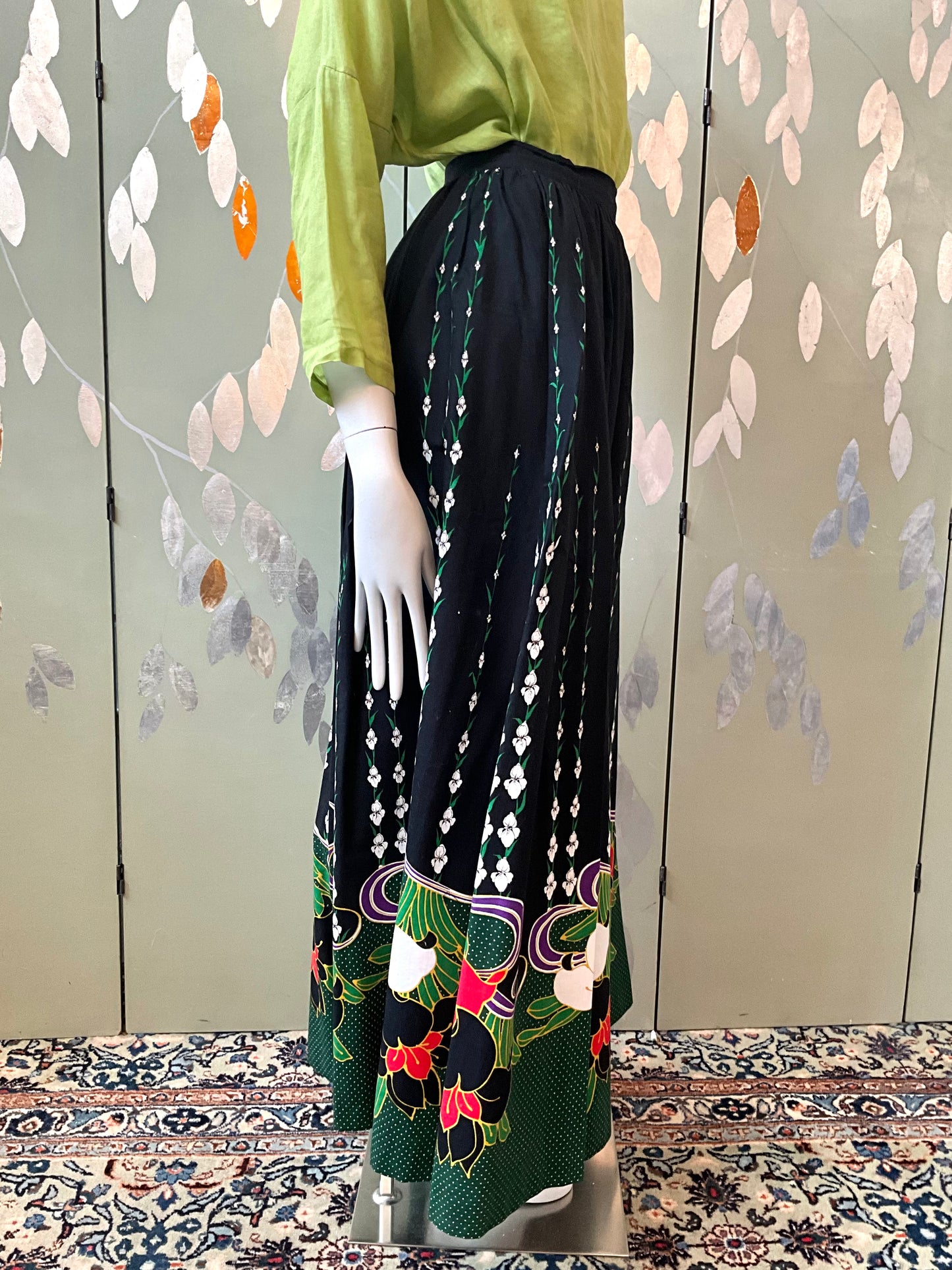 Mannequin wearing a green top and black patterned skirt against a decorative wall.