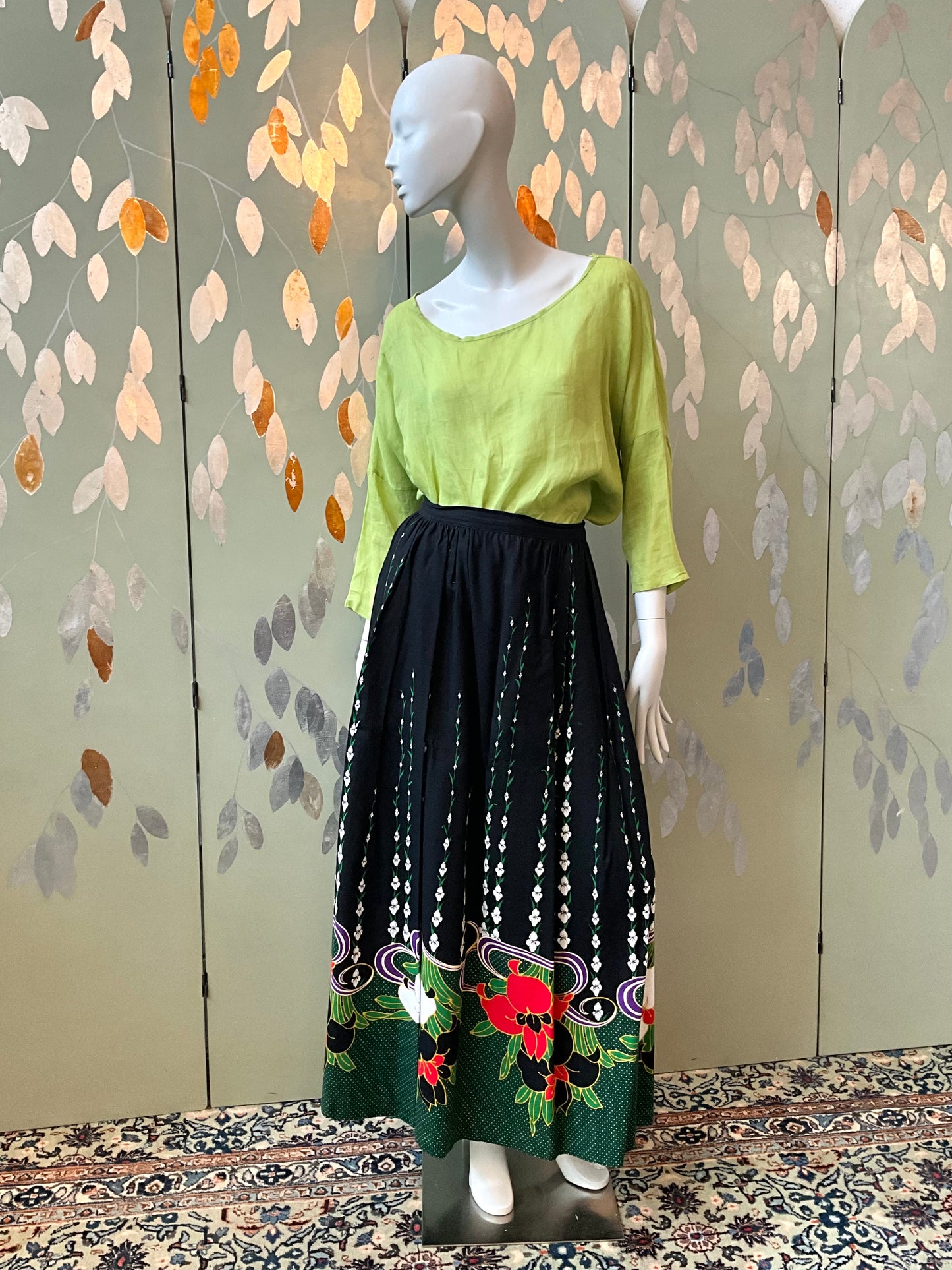 Mannequin wearing a green top and black floral skirt against a decorative wall.