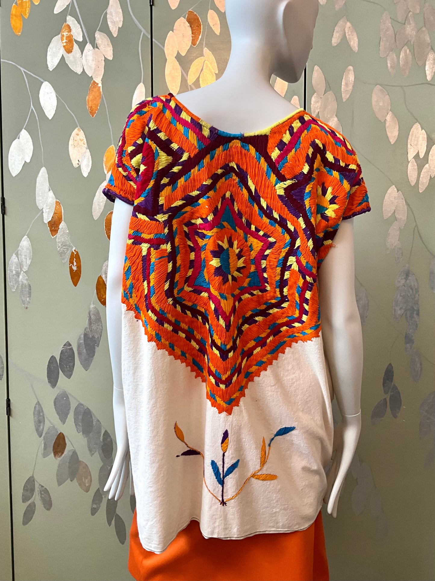 Mannequin wearing a colorful patterned top with an orange skirt against a leaf-patterned wall.