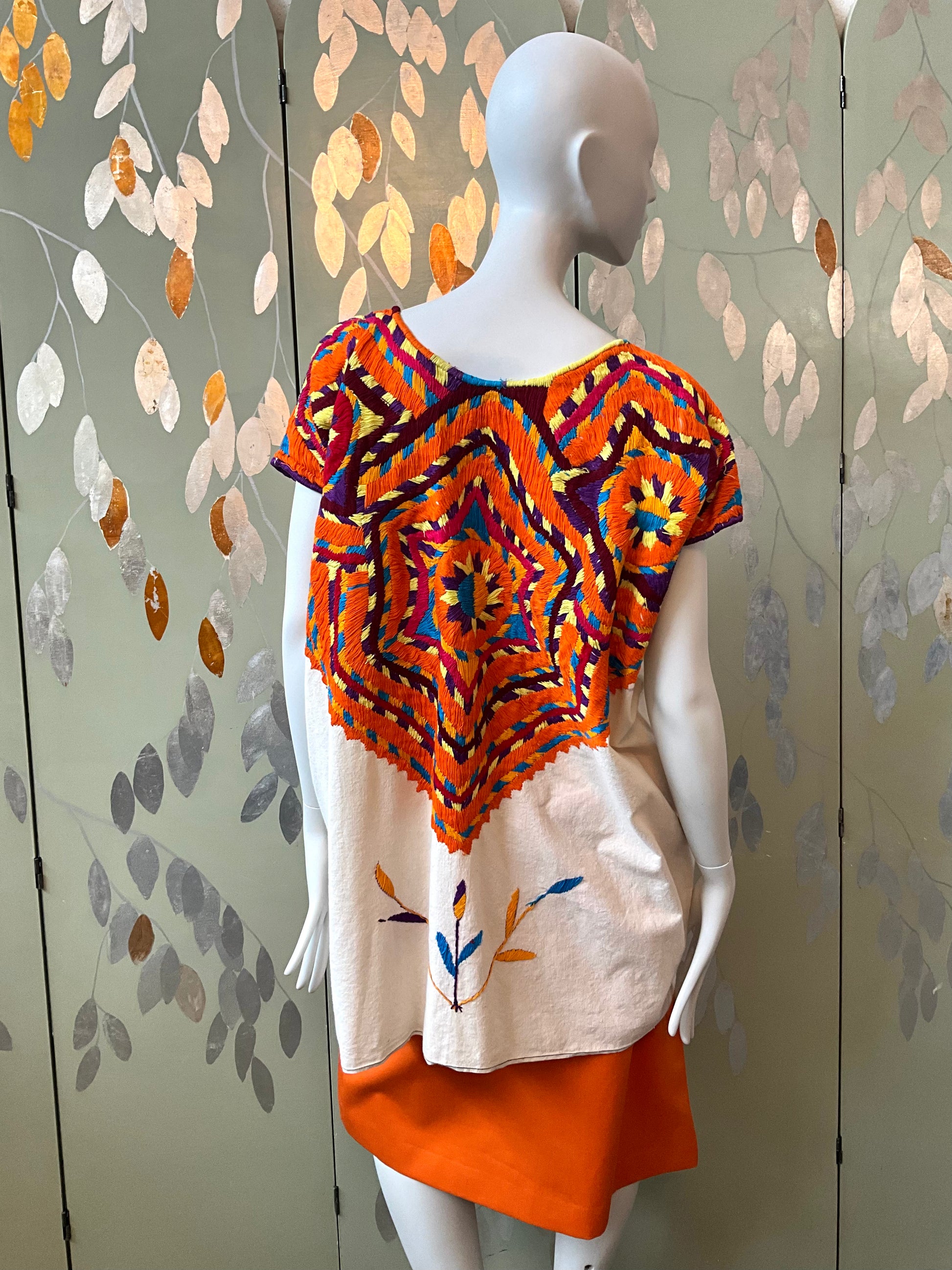 Mannequin wearing a colorful tunic with a geometric pattern against a patterned wall.