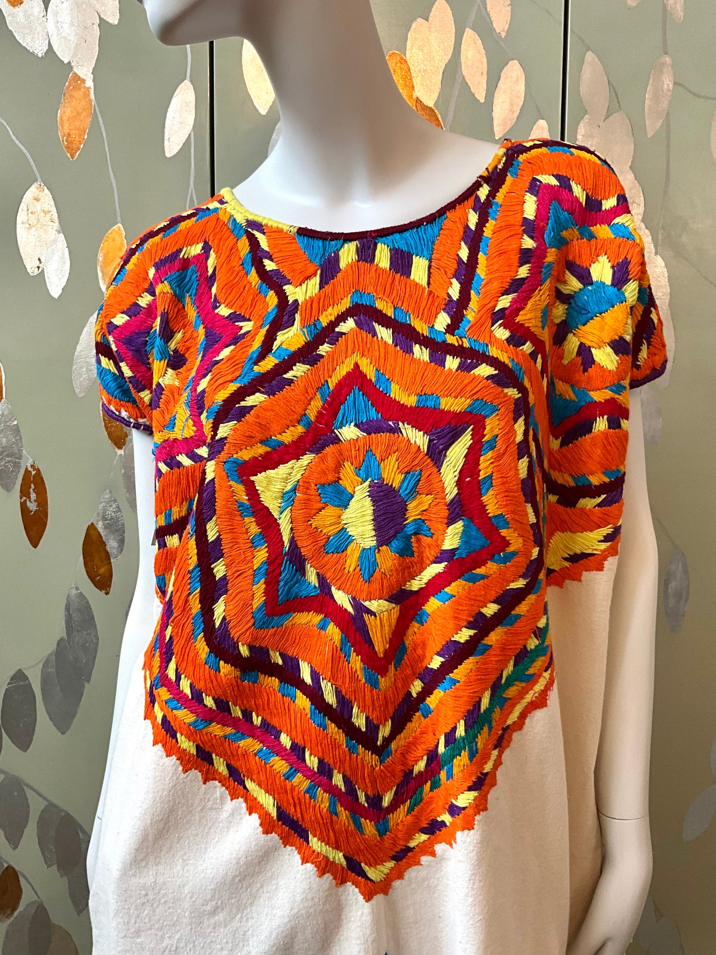 Mannequin wearing a colorful embroidered top with a geometric pattern against a neutral background.