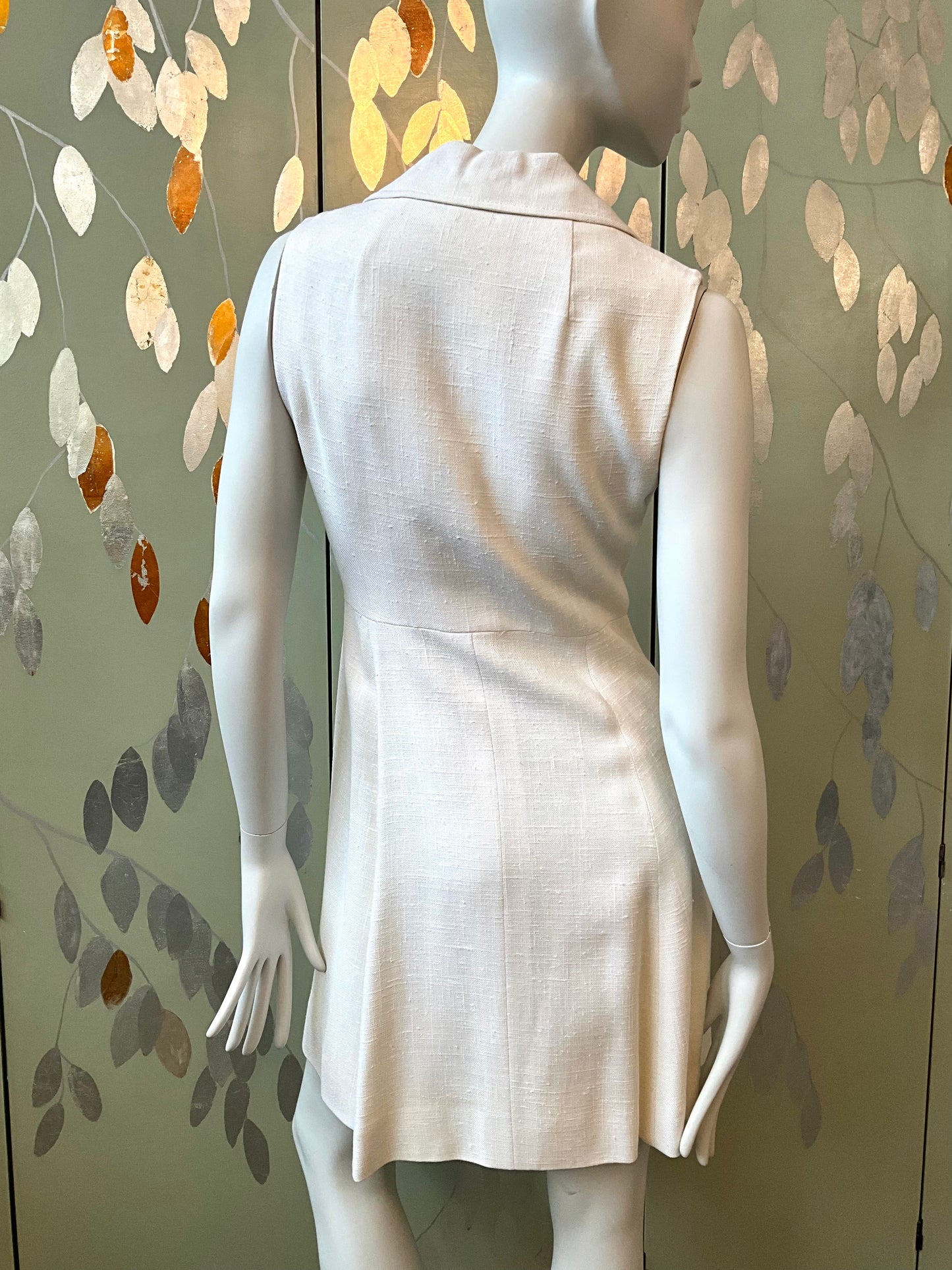 Mannequin wearing a light-colored dress against a decorative wall with leaf patterns.