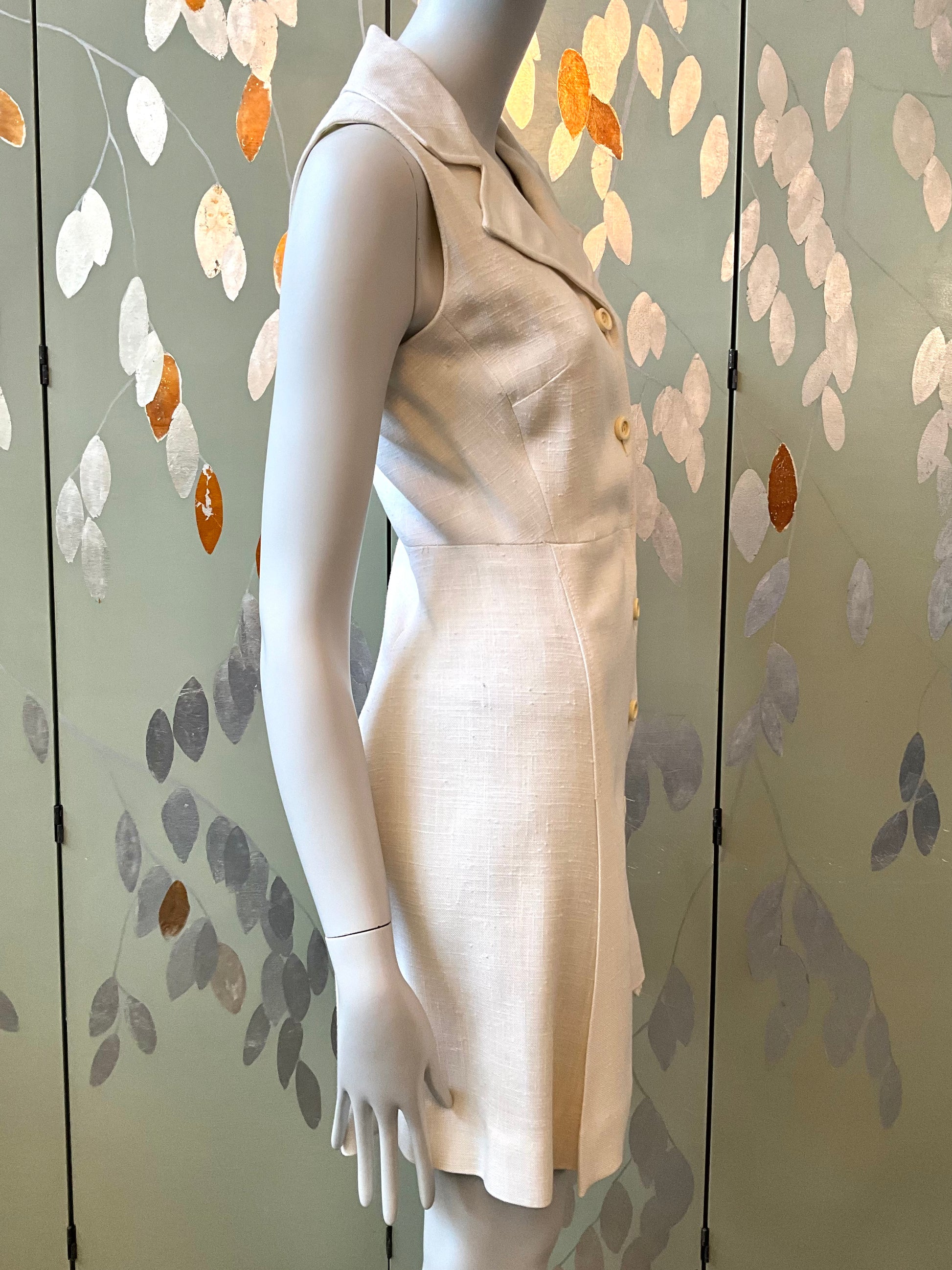 Mannequin wearing a white dress with cream buttons against a decorative wall.