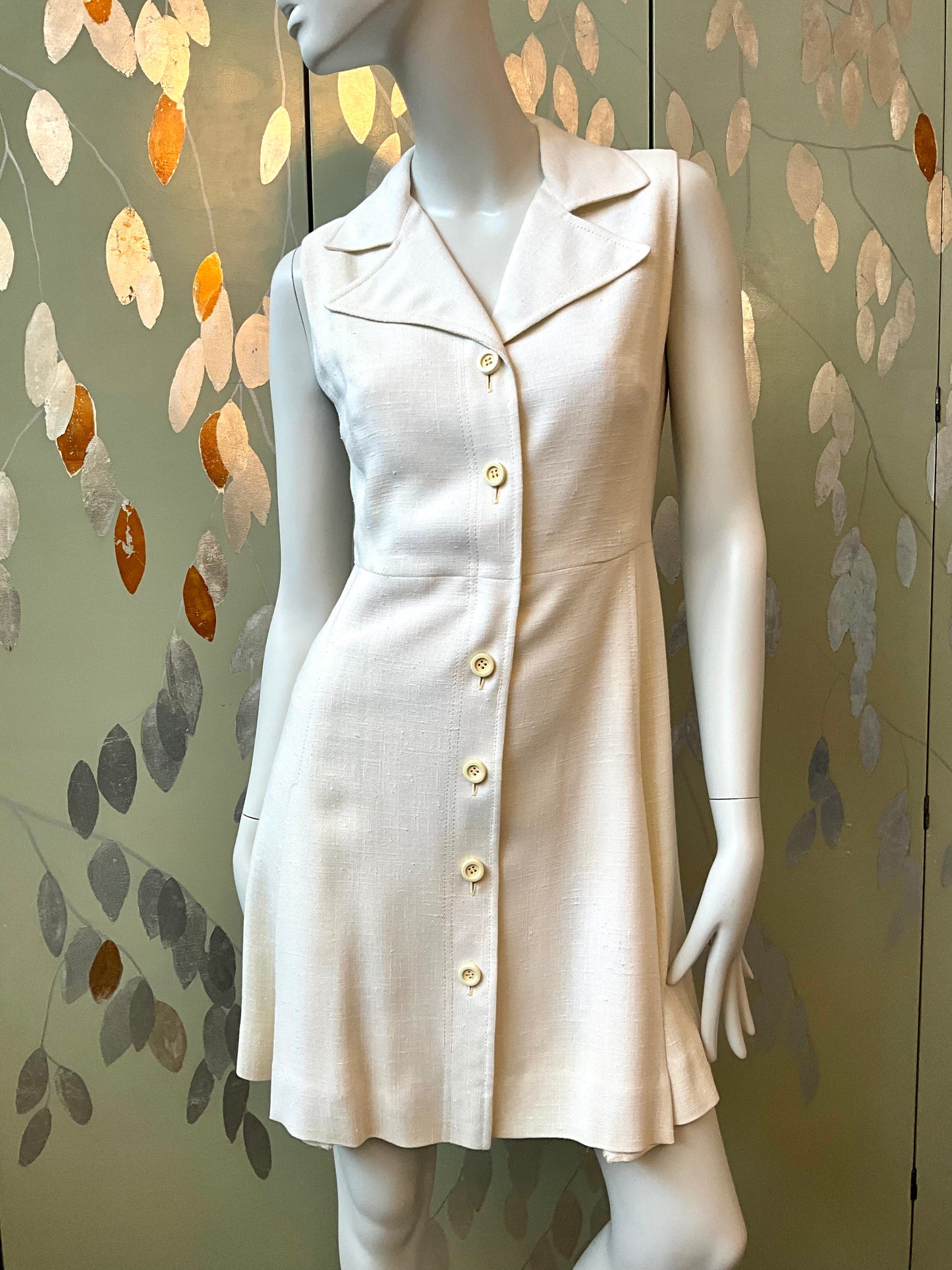 Mannequin wearing a white dress with buttons against a decorative wall.