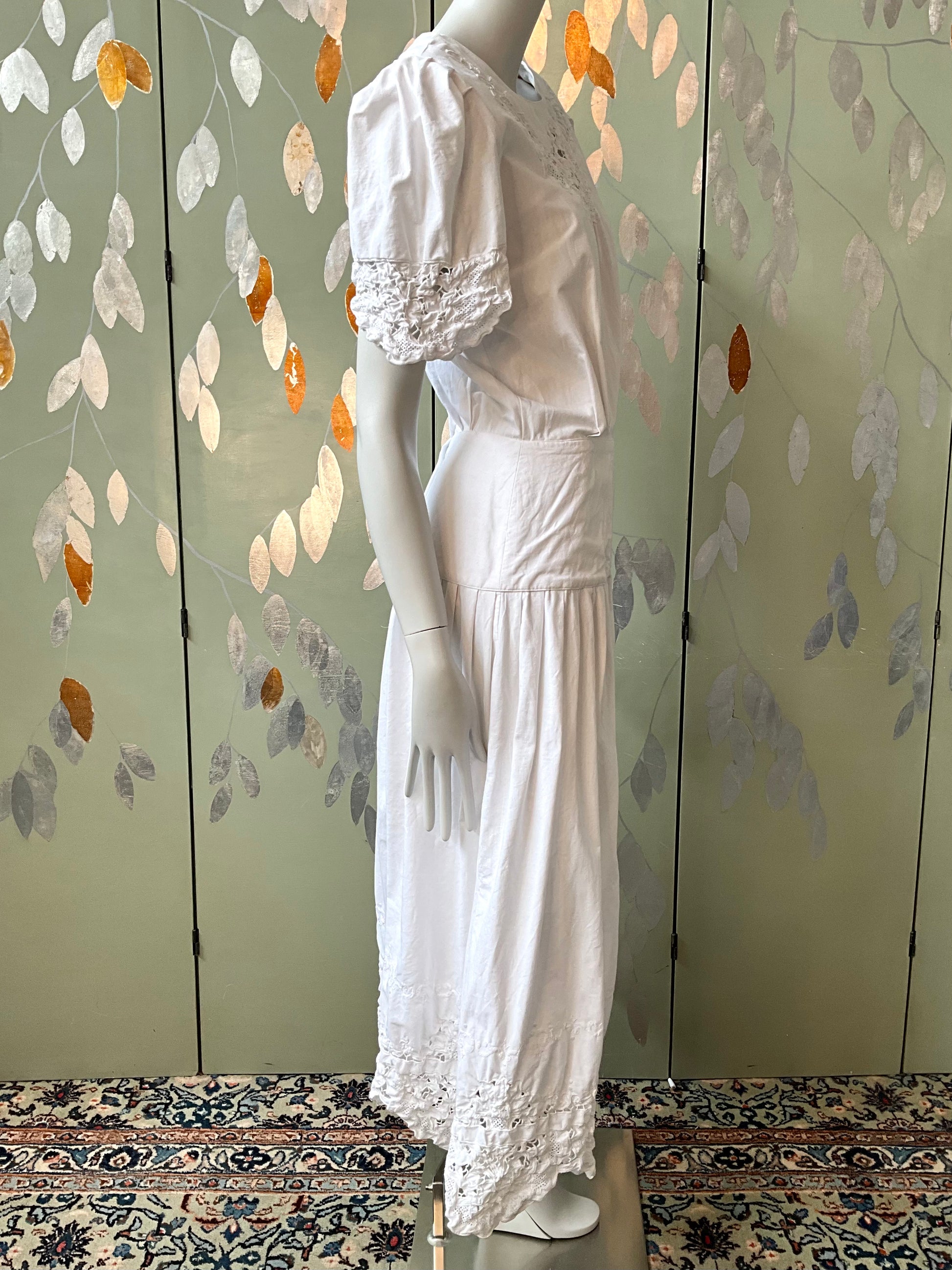 Mannequin wearing a white dress against a decorative wall with leaf patterns.