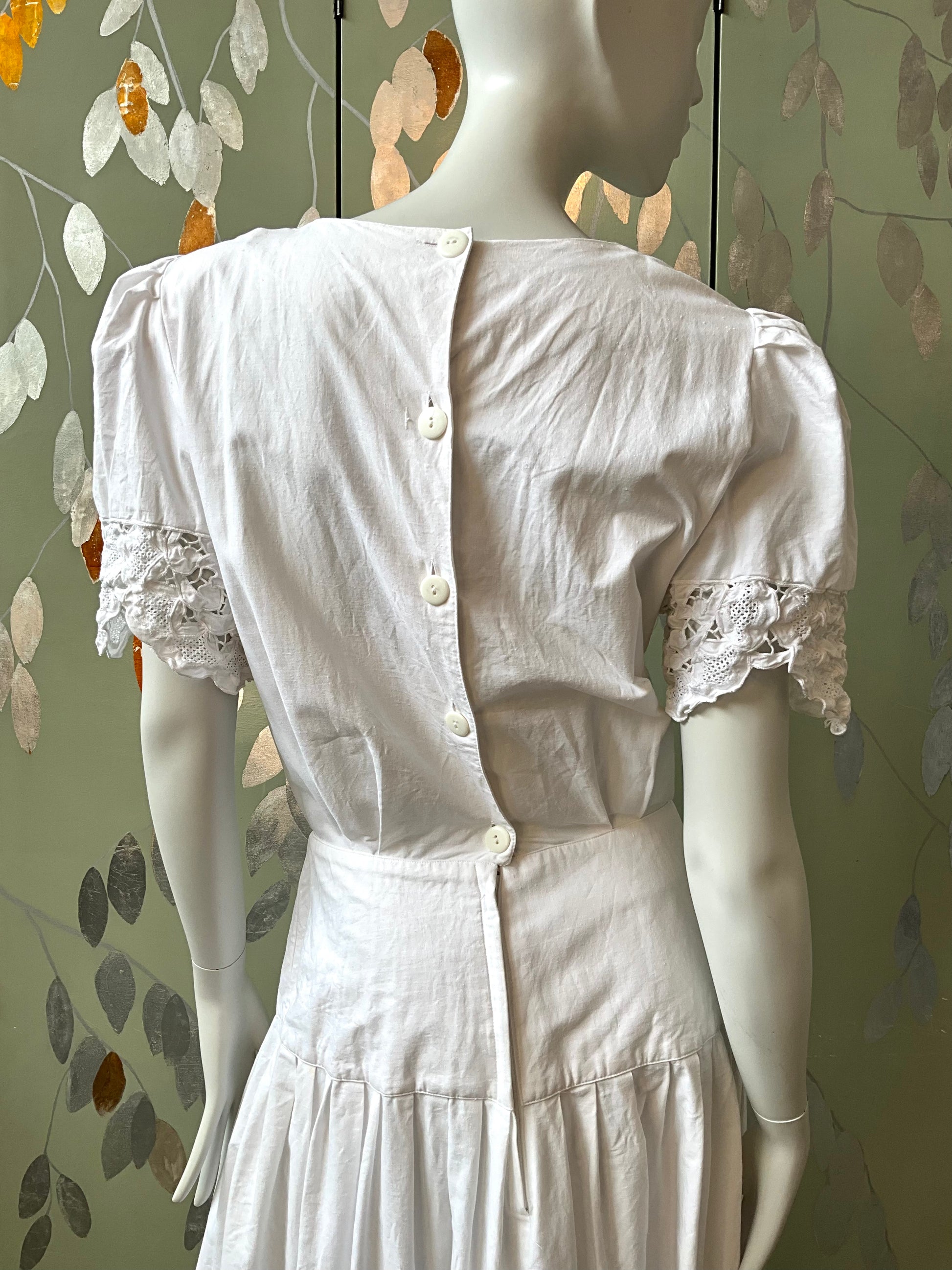 White dress with lace sleeves on a mannequin against a leaf-patterned wall.