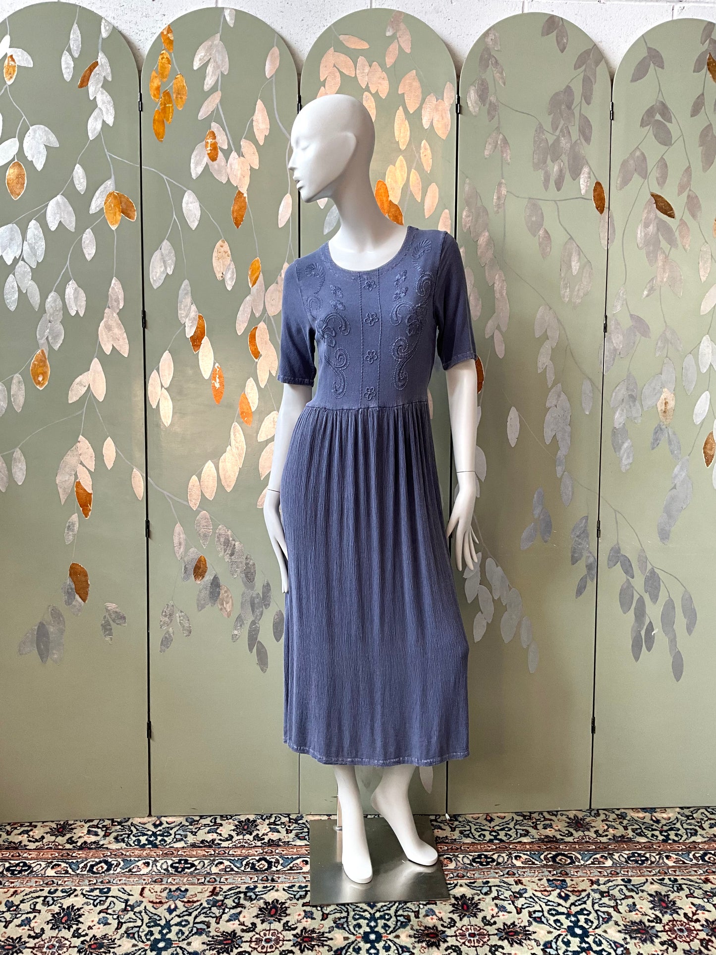 Mannequin wearing a blue dress against a decorative wall with leaf patterns.