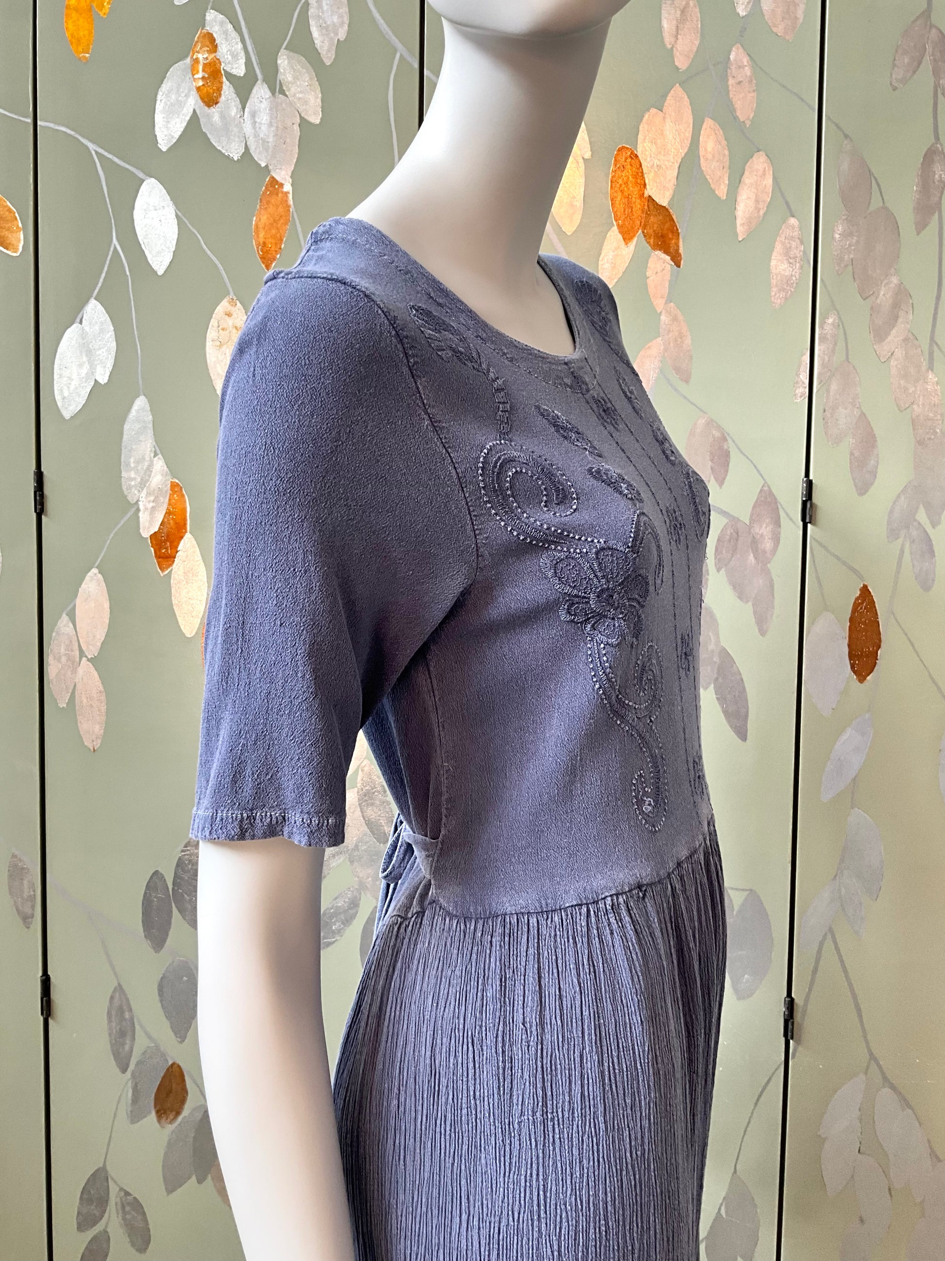 Mannequin wearing a blue dress with floral embroidery against a decorative wall.