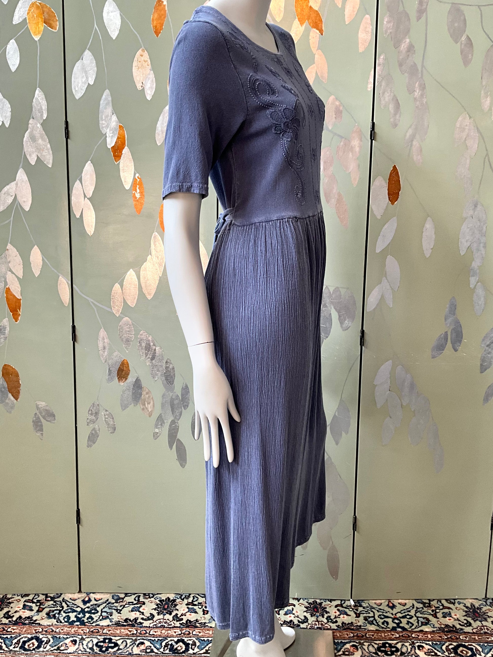 Mannequin wearing a blue dress against a decorative wall with leaf patterns.