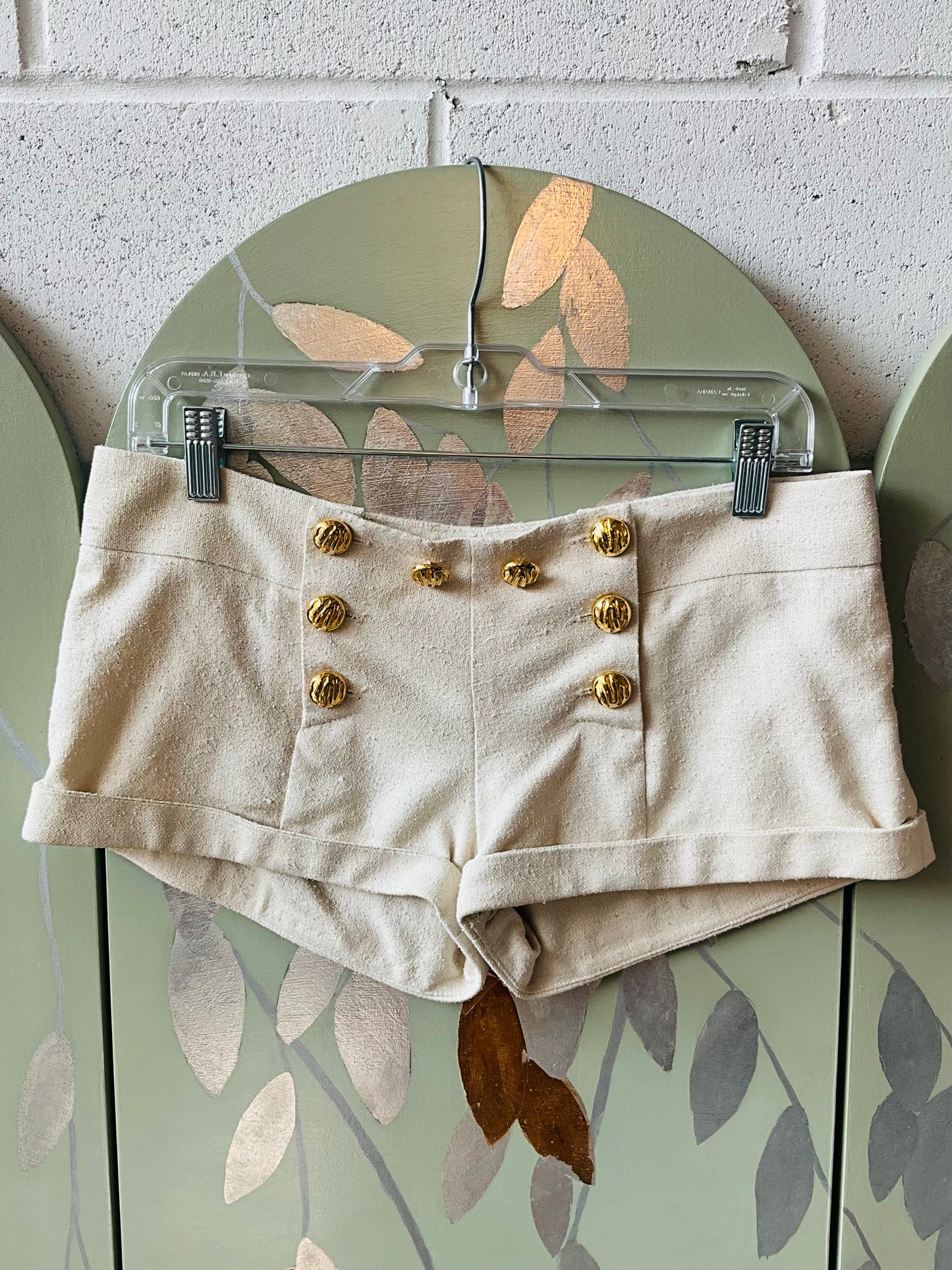 Beige shorts with gold buttons on a hanger against a wall.