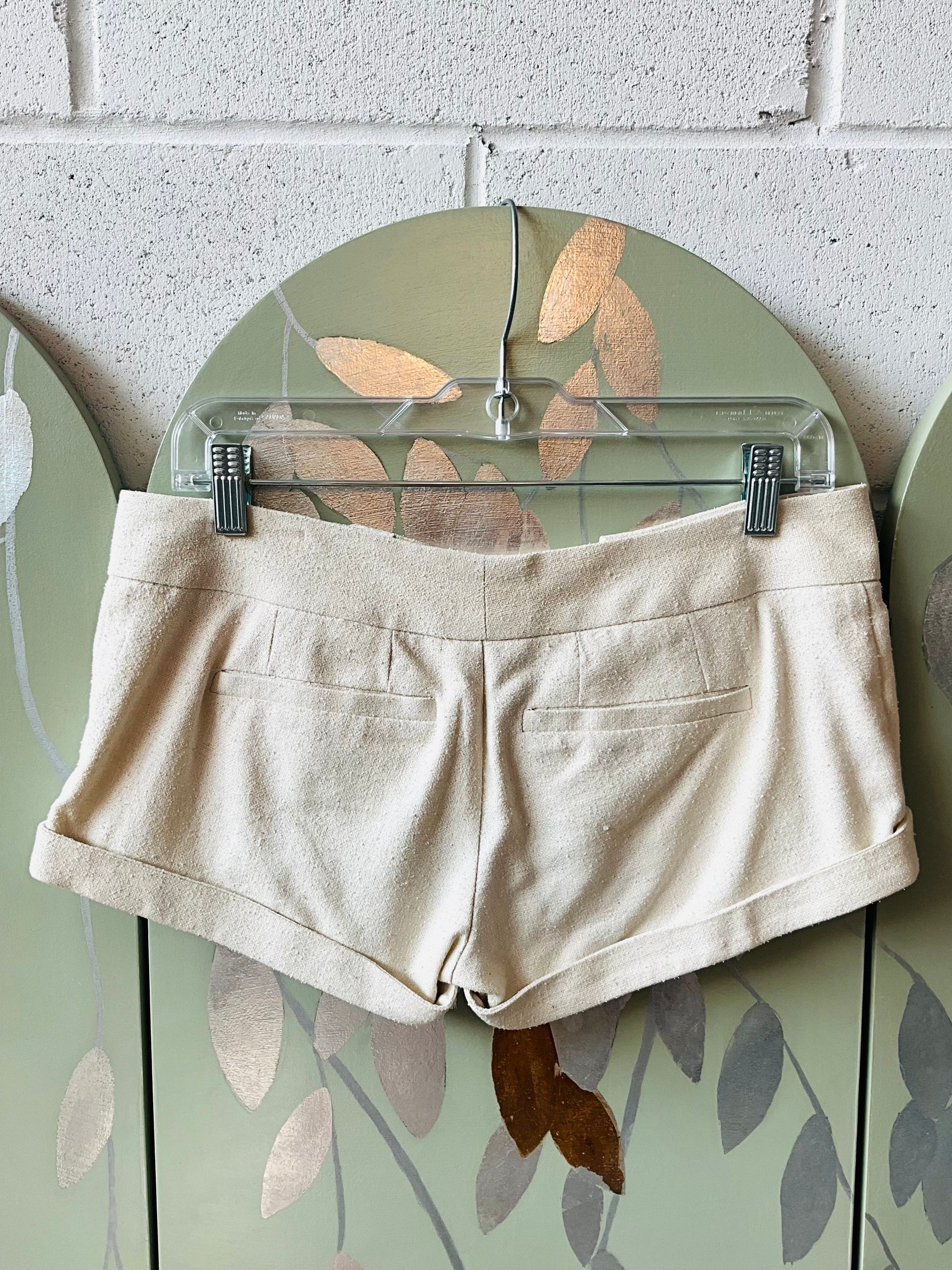 Beige shorts on a hanger against a decorative wall with leaf patterns.
