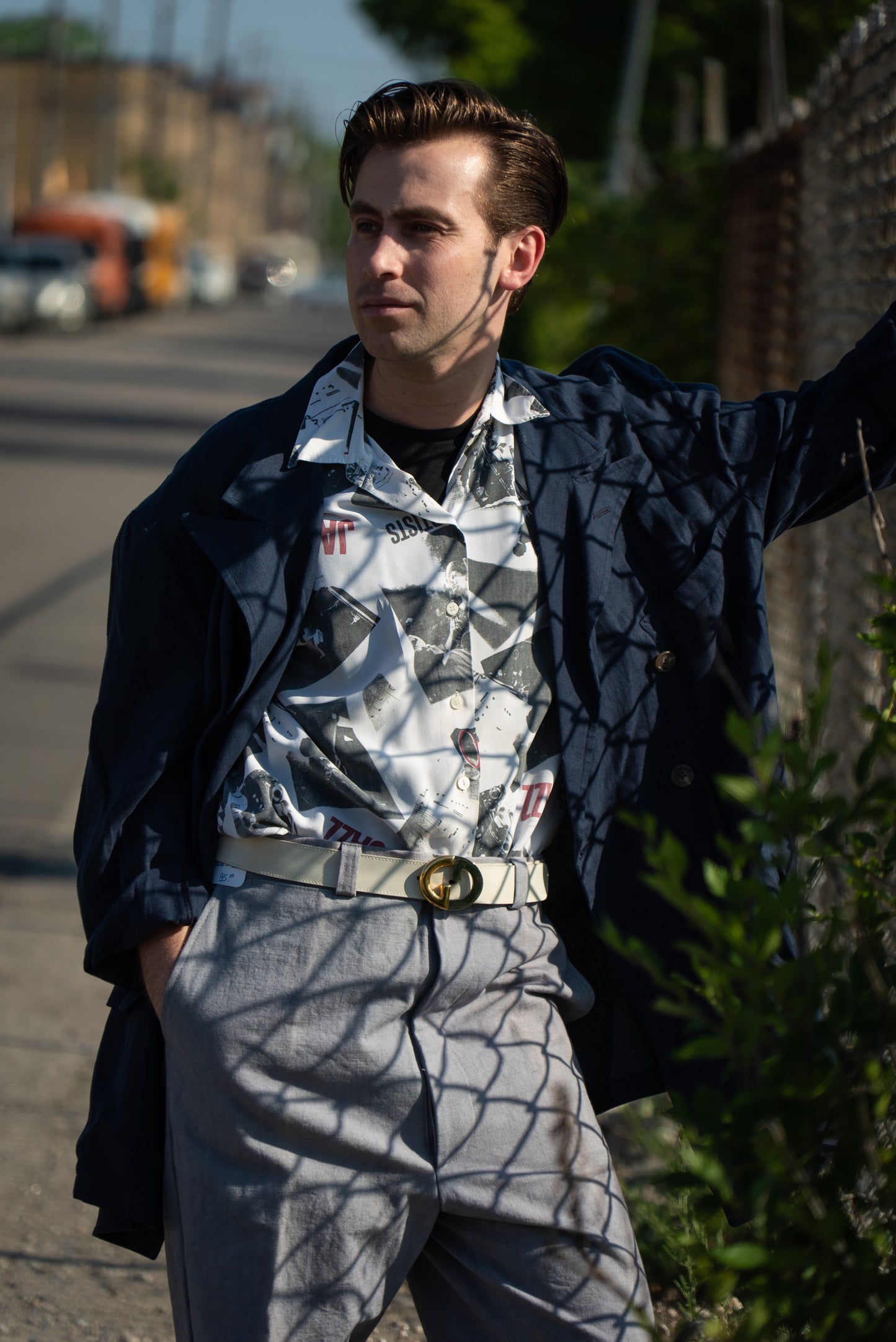 Man wearing a patterned shirt and gray pants standing outdoors with a blurred street background