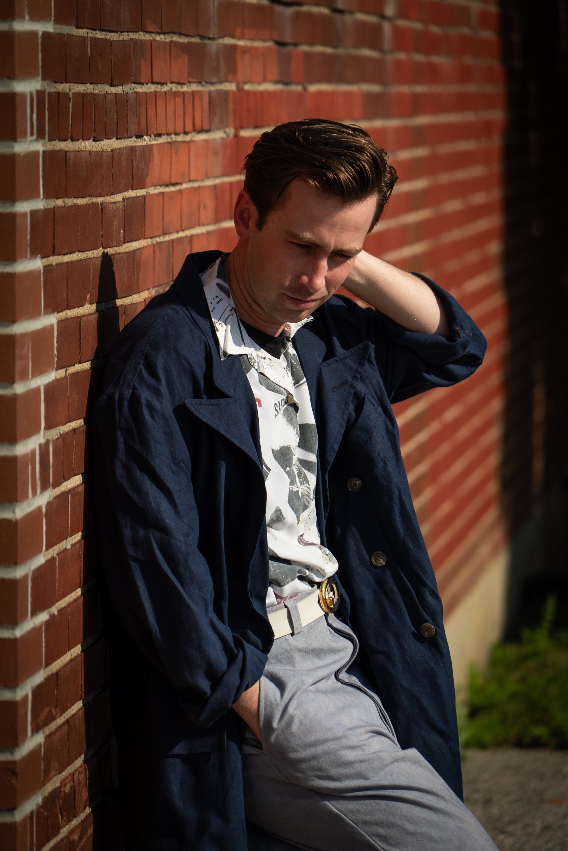 Man leaning against a brick wall wearing a navy jacket and gray pants.