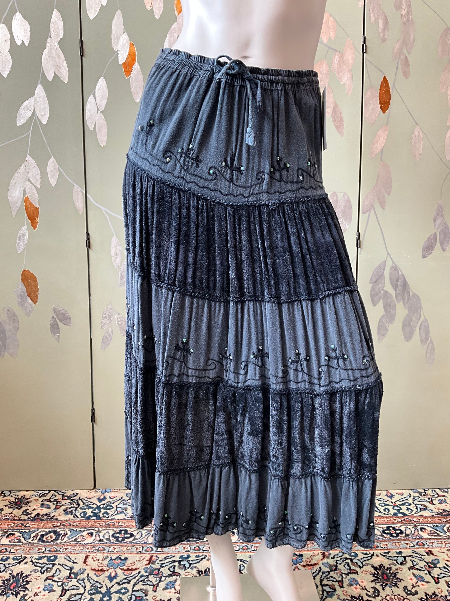 Long grey skirt with layered design on a mannequin against a floral-patterned wall.