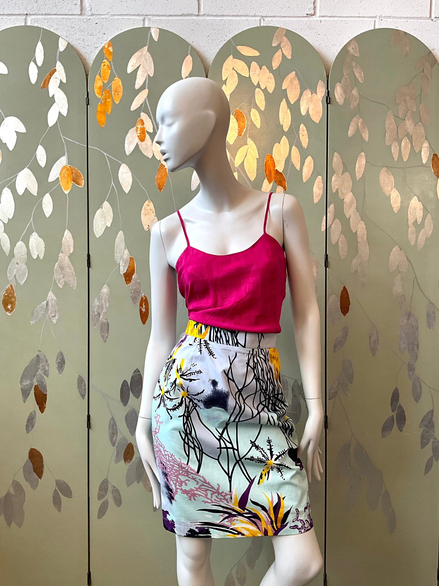 Mannequin wearing a pink top and patterned skirt against a decorative screen with leaf design.