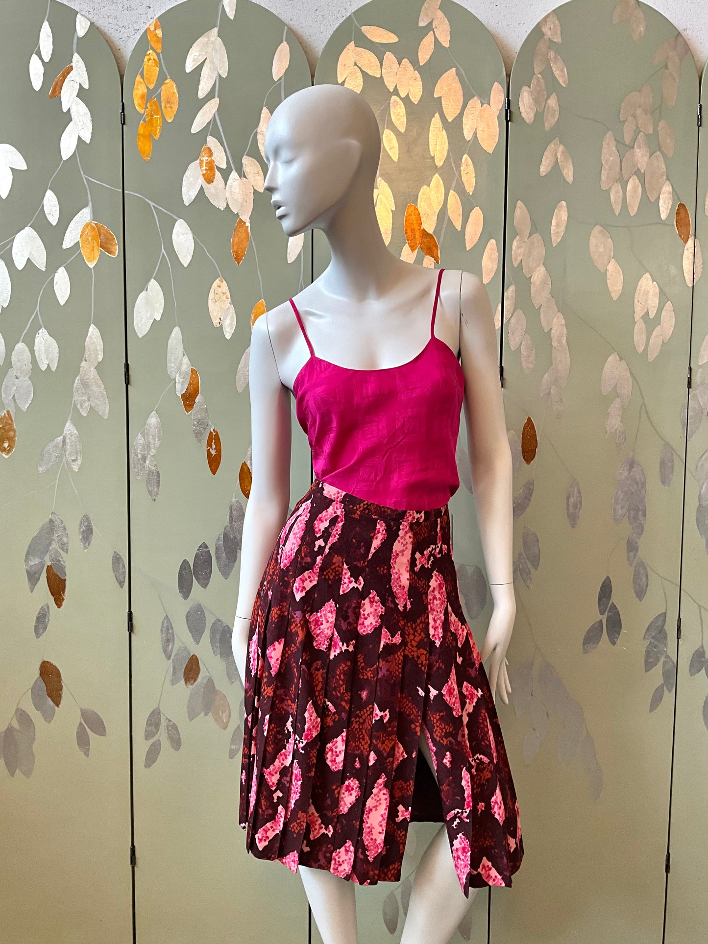 Mannequin wearing a pink top and patterned skirt against a decorative screen with leaf designs.