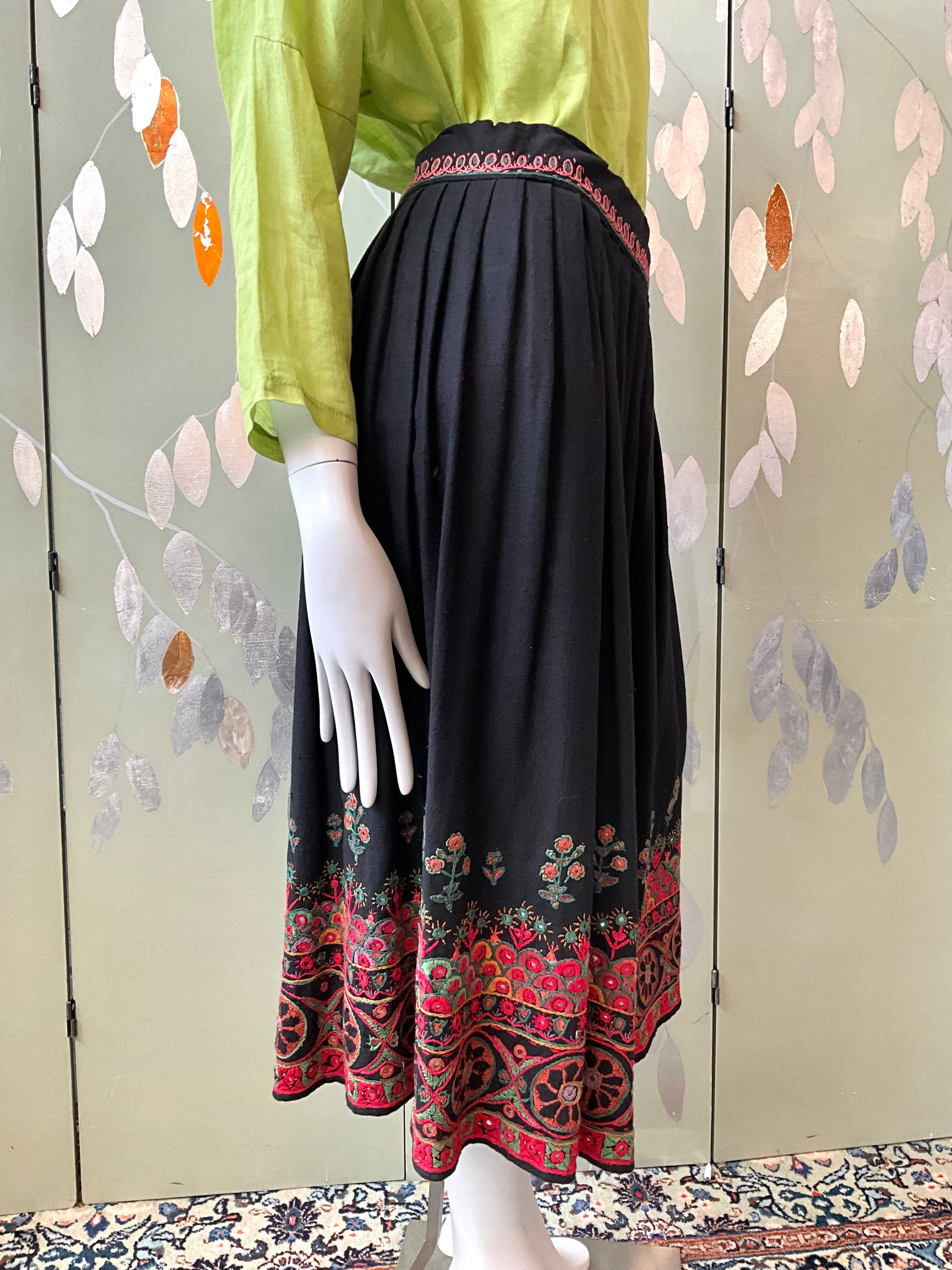 Mannequin wearing a green top and black skirt with red patterns against a floral-patterned wall.