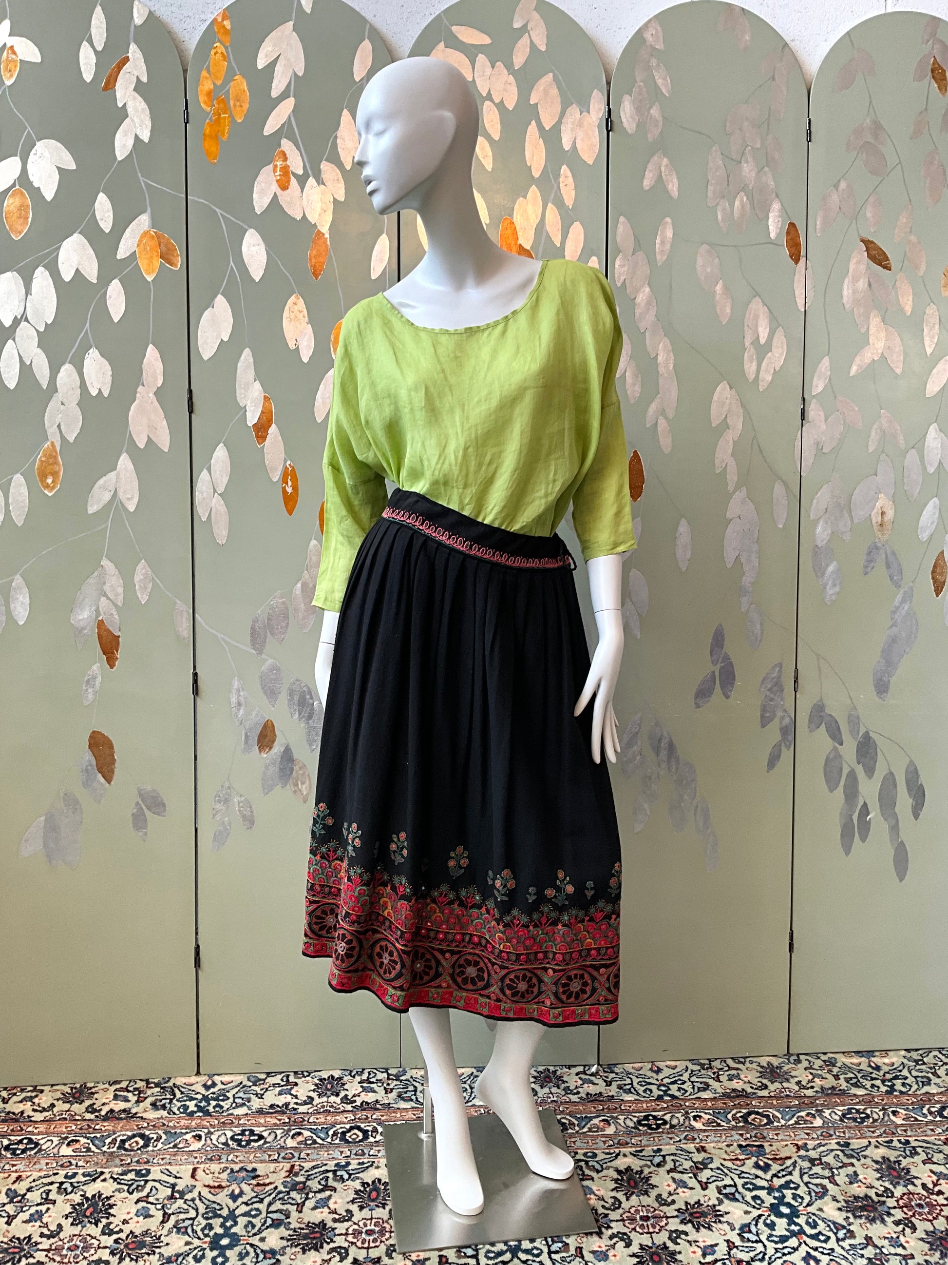 Mannequin wearing a green top and black skirt with red patterns against a decorative wall.