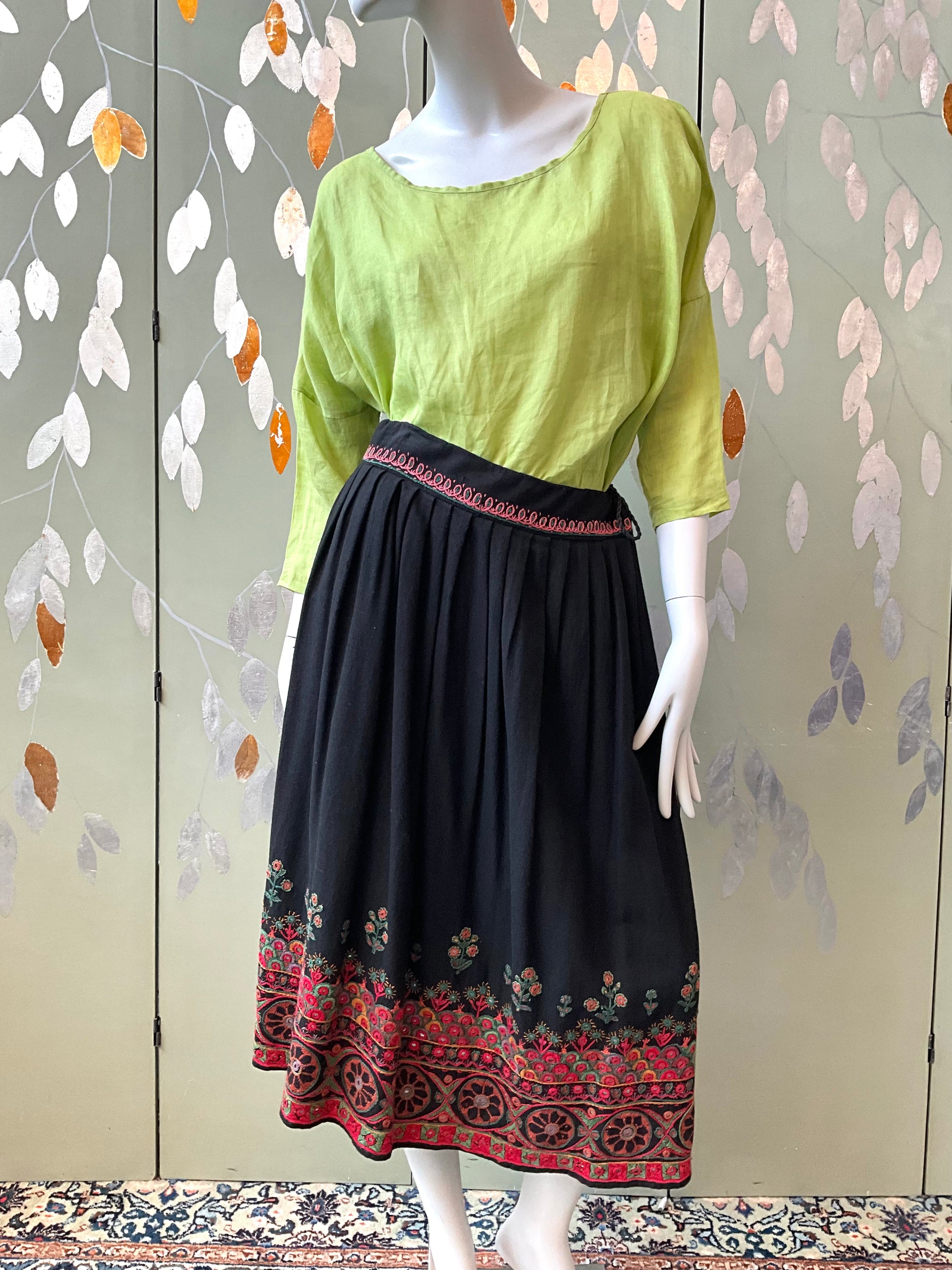 Mannequin wearing a green top and black skirt with red patterns against a decorative wall.