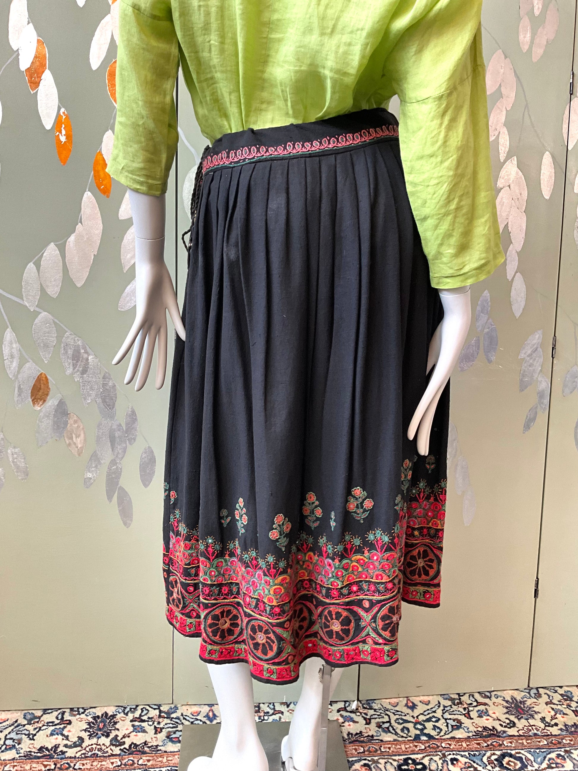 Mannequin wearing a black skirt with red and gold patterns and a green top against a decorative wall.