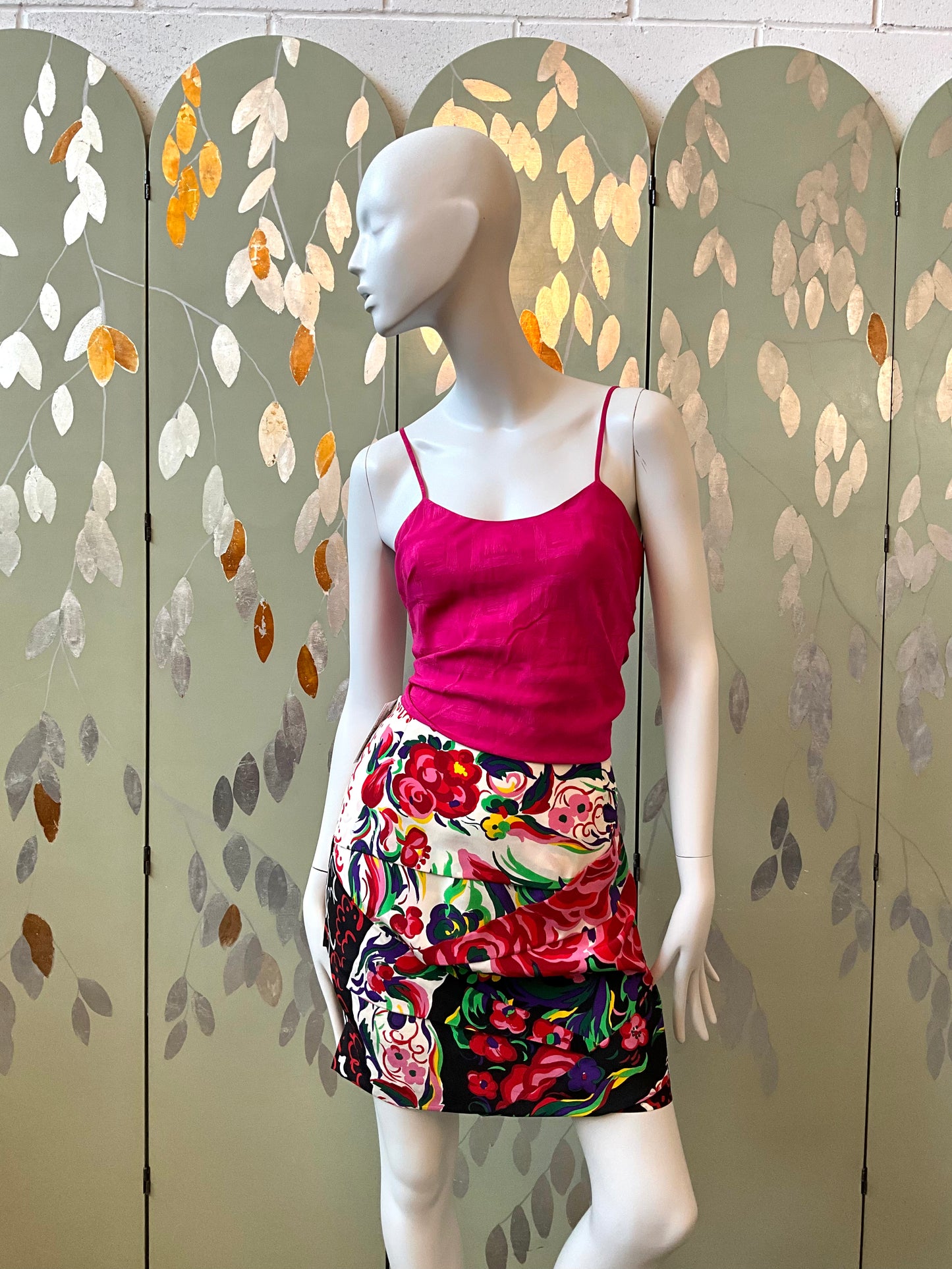 Mannequin wearing a pink top and floral skirt against a decorative screen with leaf patterns.